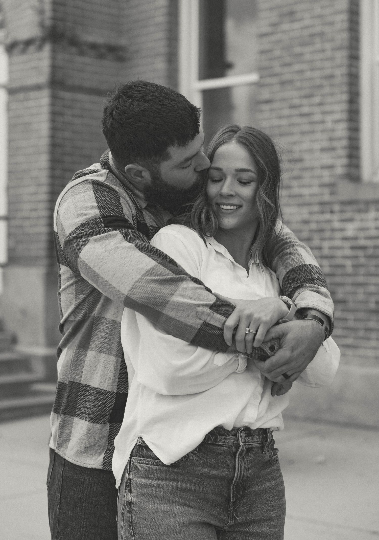 A romantic black and white portrait of a couple embracing against a brick building background.