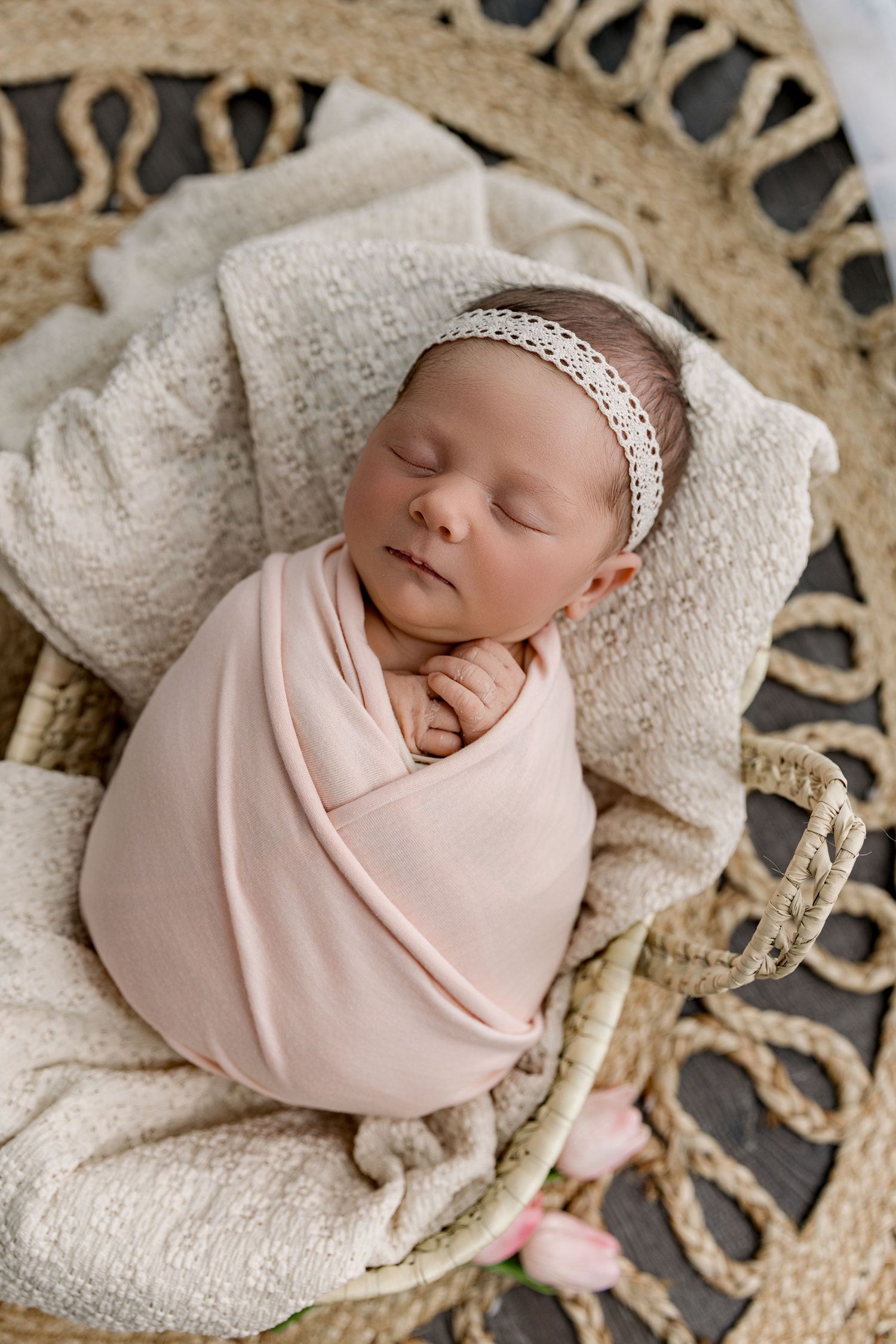Sleeping newborn baby girl swaddled in pink wrap with lace headband resting in a wicker basket.