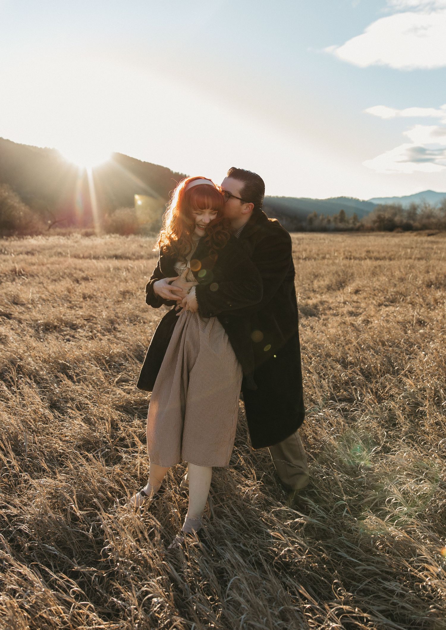 Couple sharing intimate moments in golden sunlit field during sunset with mountains in background.