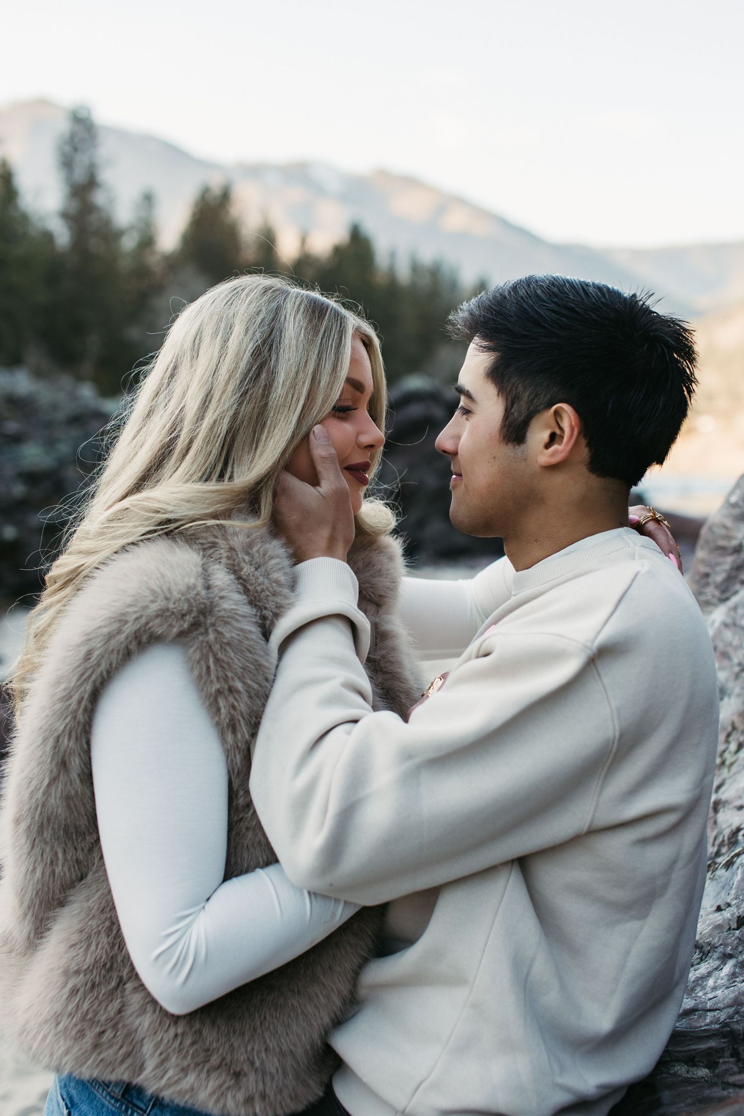 A romantic couple in cream colored outfits share tender moments against a mountain backdrop during sunset.
