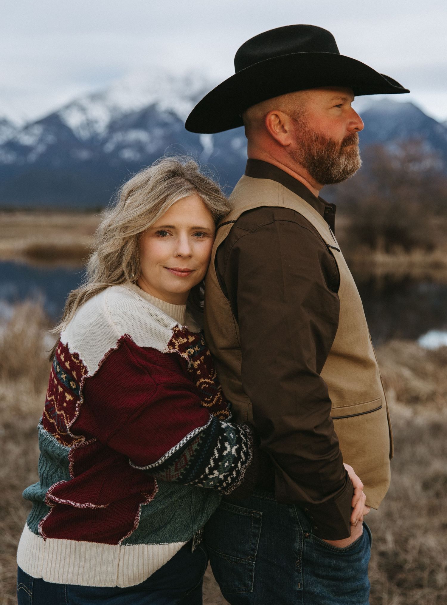 A couple in western attire embraces against a backdrop of snow-capped mountains in a romantic outdoor photoshoot.