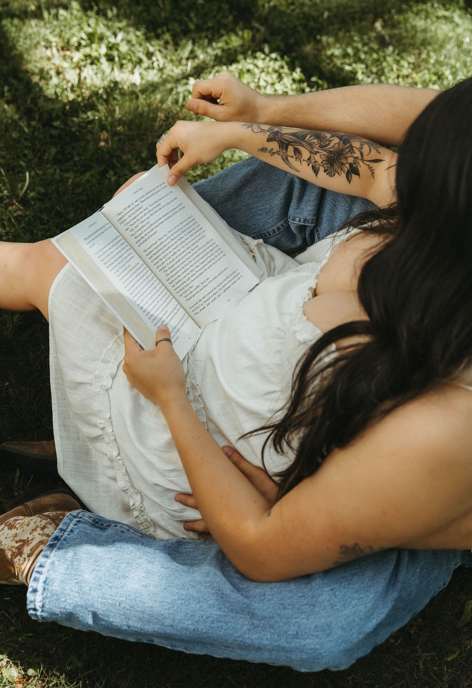 A person in a white dress lying on a blue blanket reads a book while relaxing outdoors in dappled sunlight.