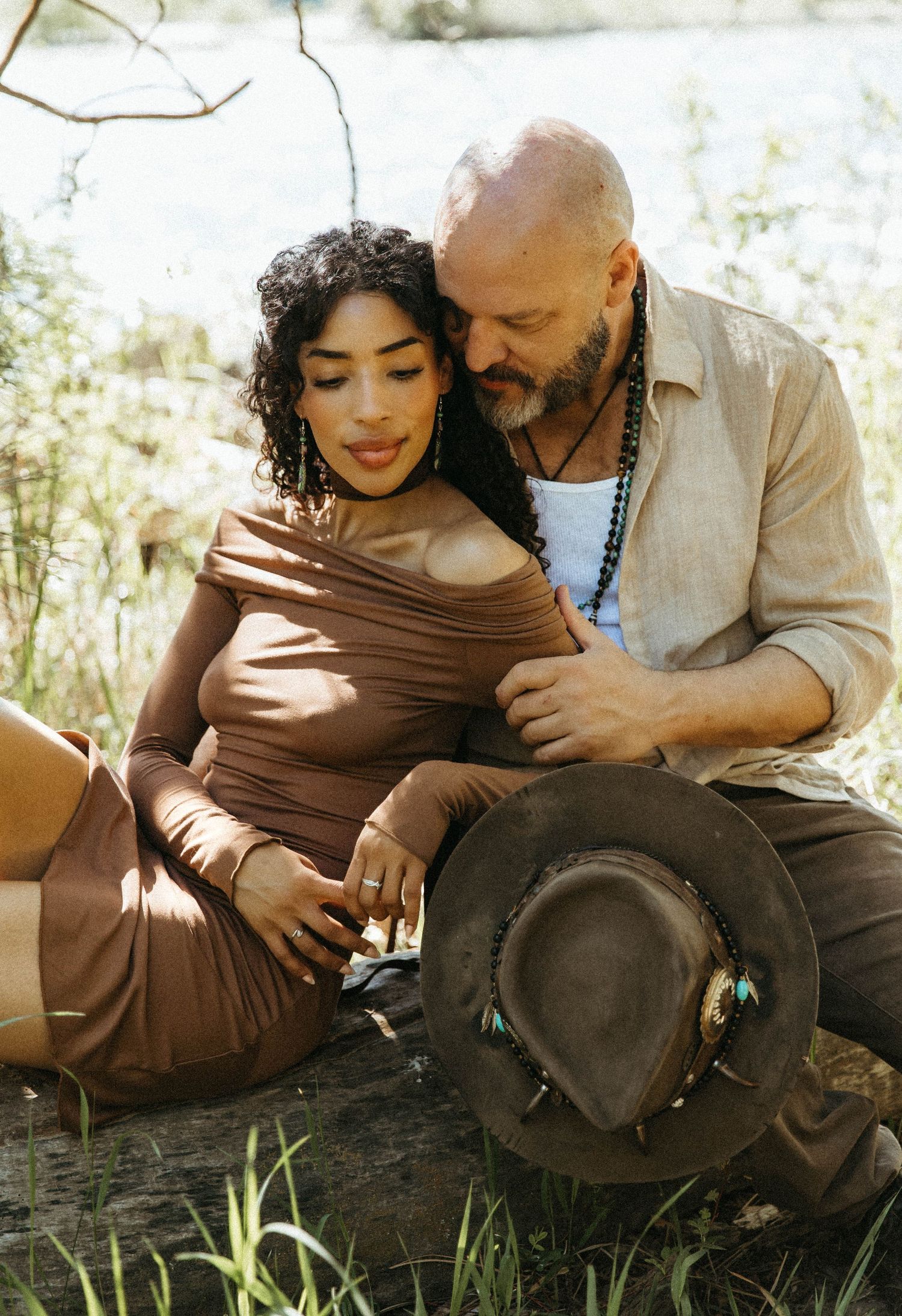 Romantic couple sitting together outdoors in sunlight with a brown hat nearby, sharing an intimate moment.