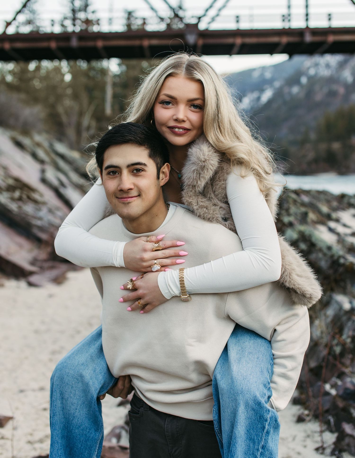 A couple in cream sweaters and jeans embracing on a rocky beach with mountains in the background.