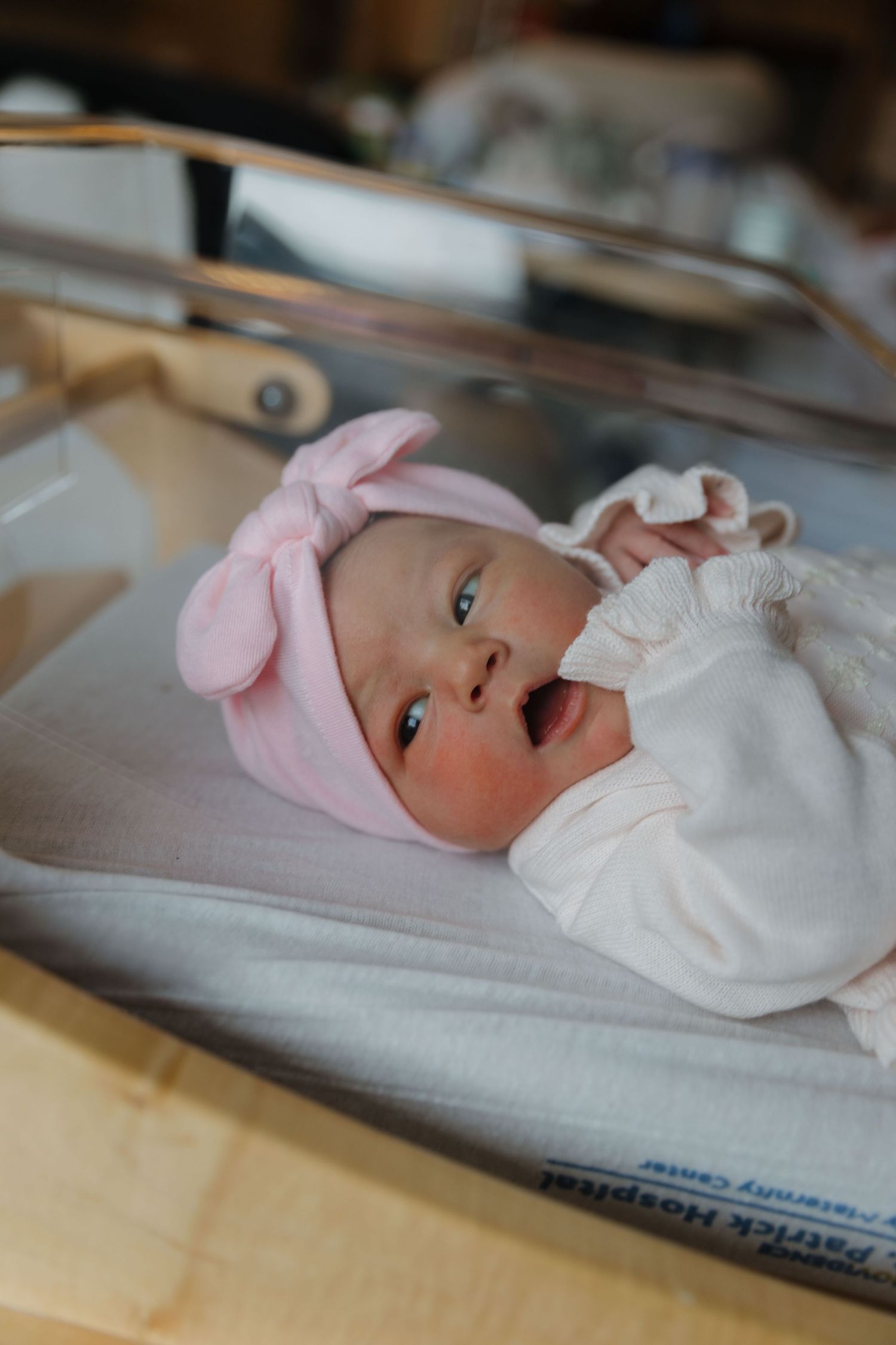 Newborn wearing pink bow headband and white outfit yawning on hospital bedding.