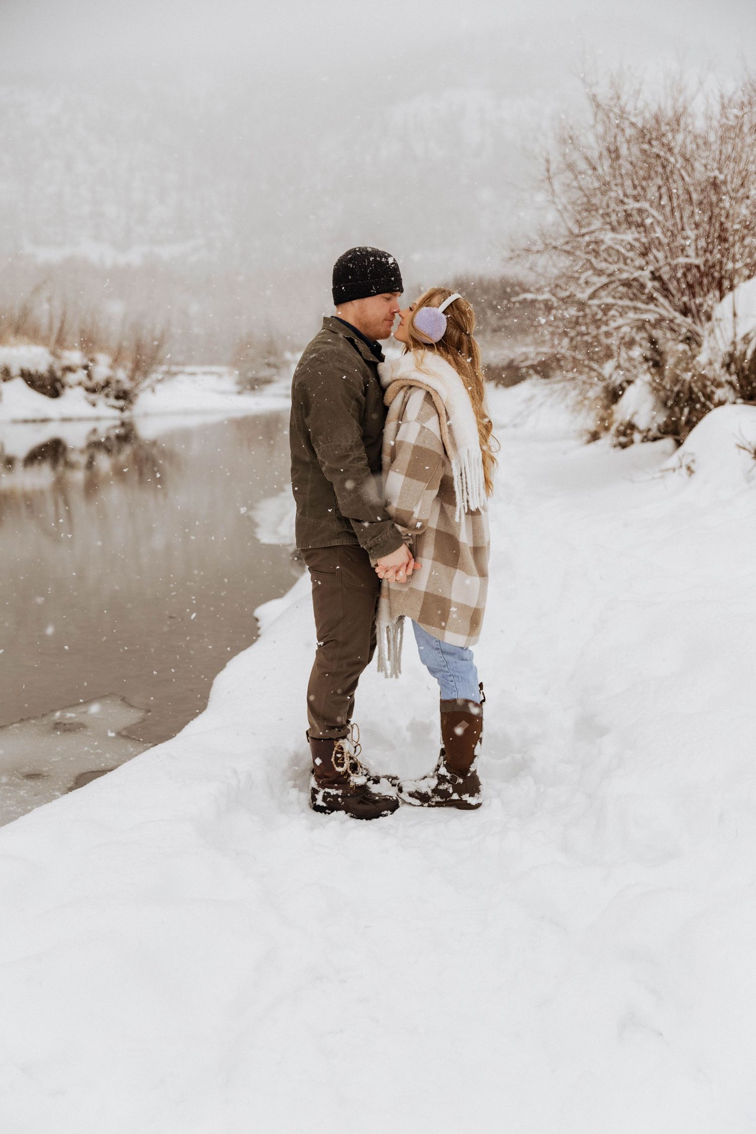 A couple stands together on a snowy riverbank during winter.
