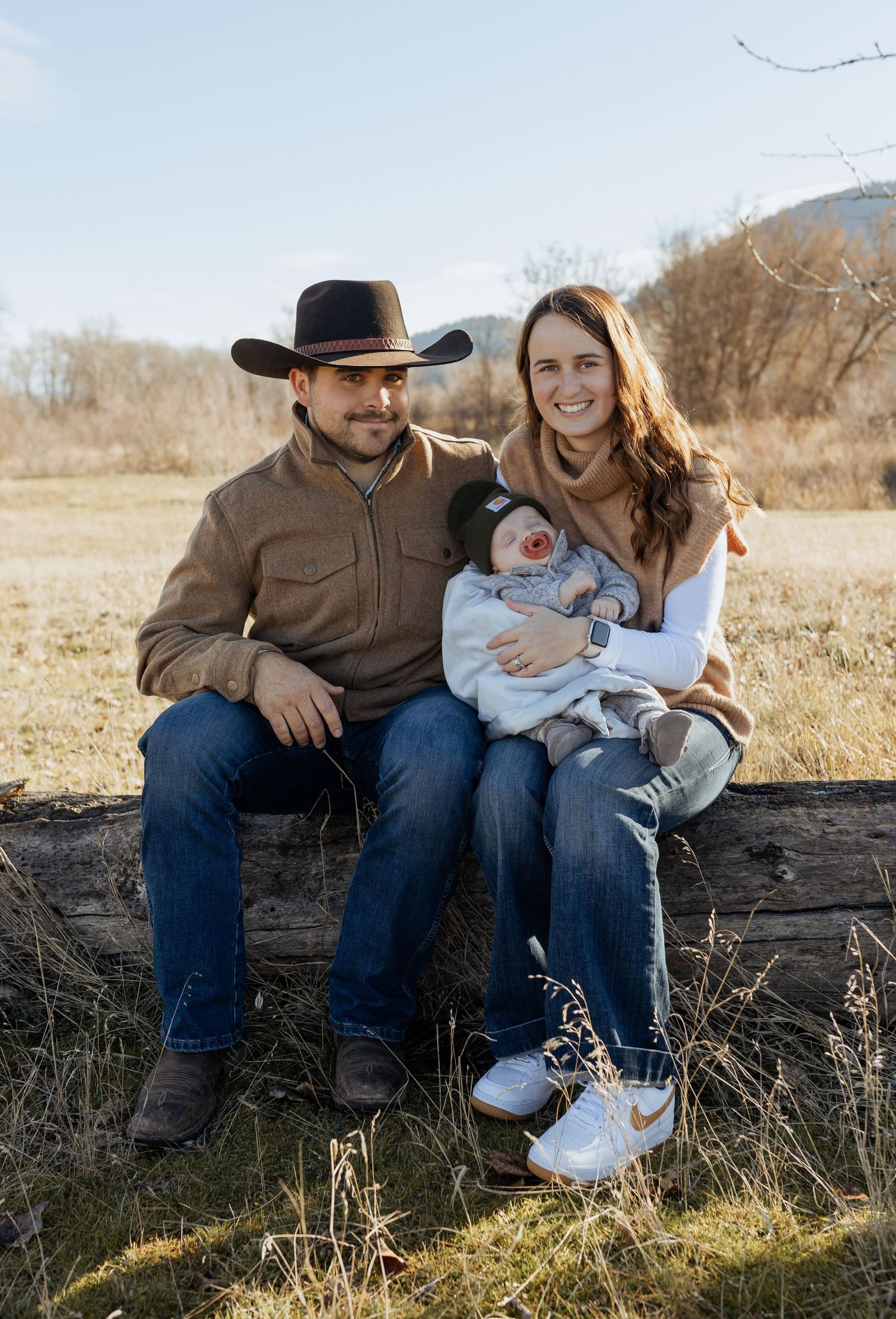 A family sits together on a fallen log in a rustic outdoor setting with mountains in the distance.