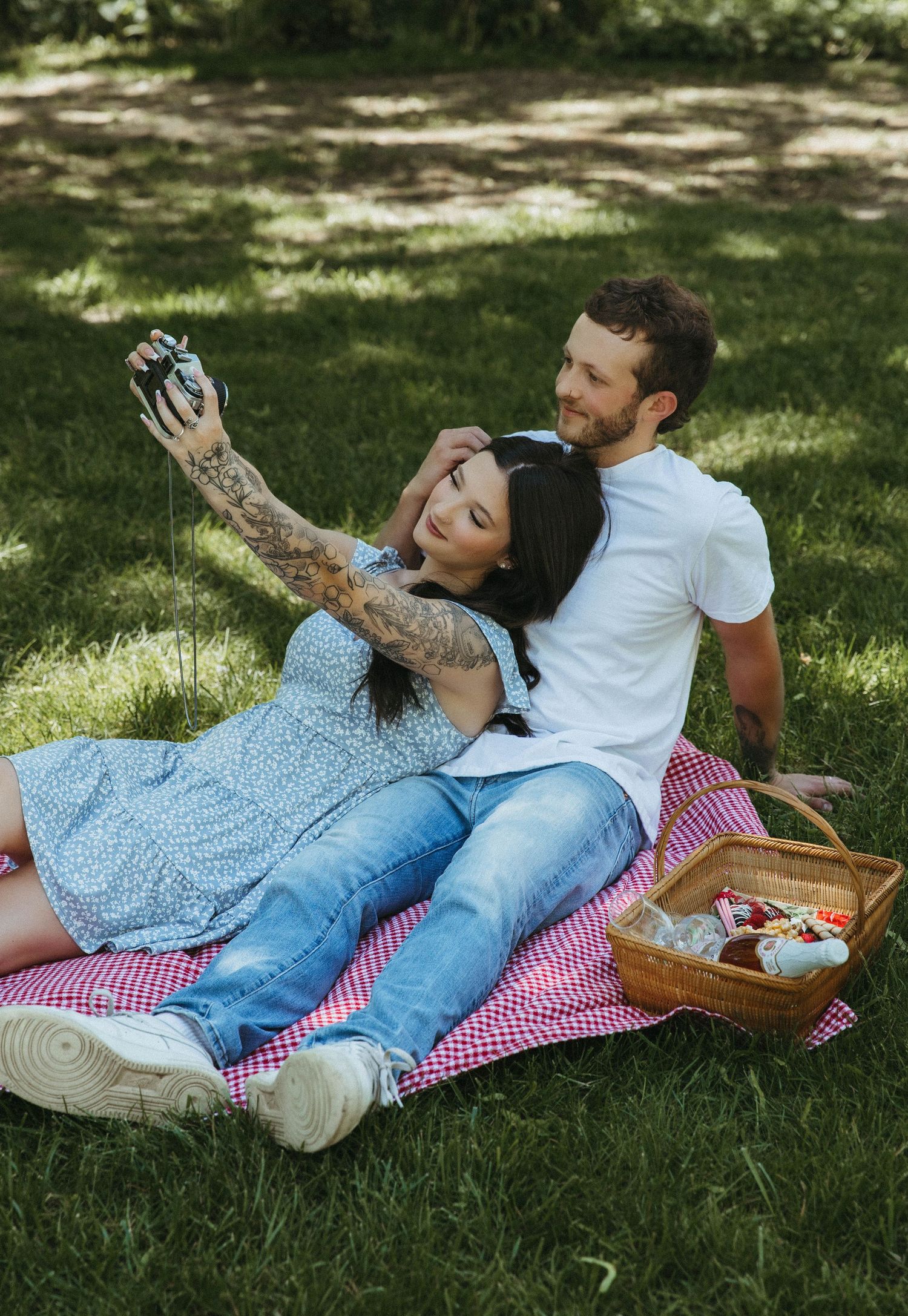 A couple relaxes together on a red and white checkered picnic blanket in a sunlit grassy park setting.