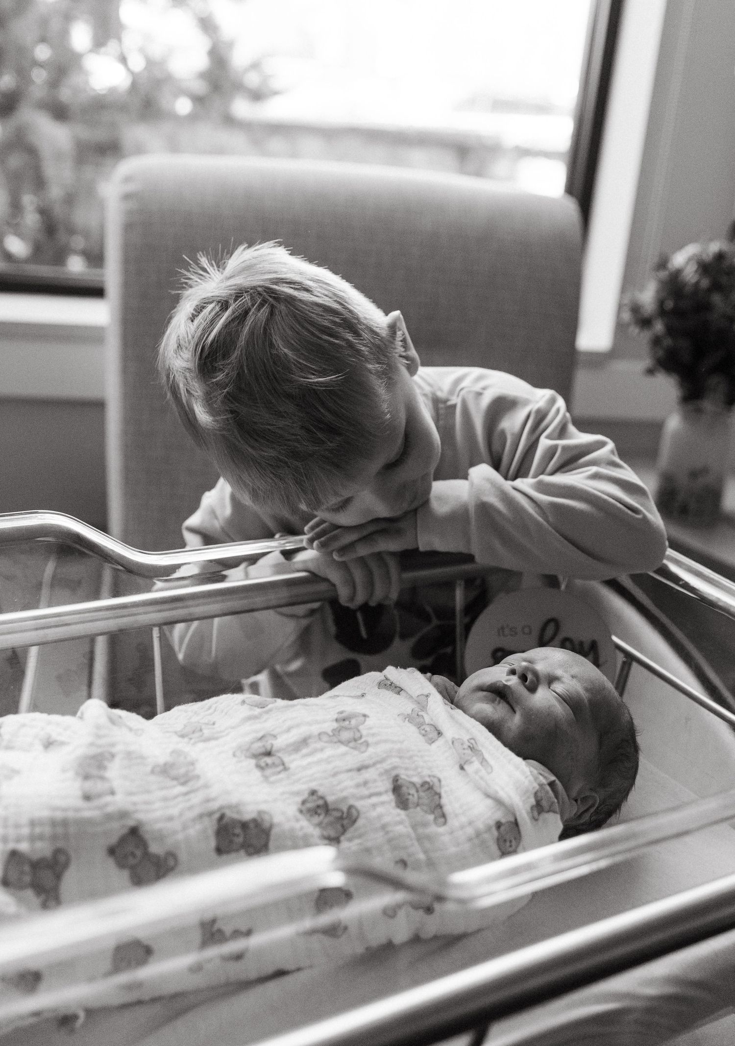 Black and white photo of a young boy leaning over hospital bassinet to kiss his newborn sibling.
