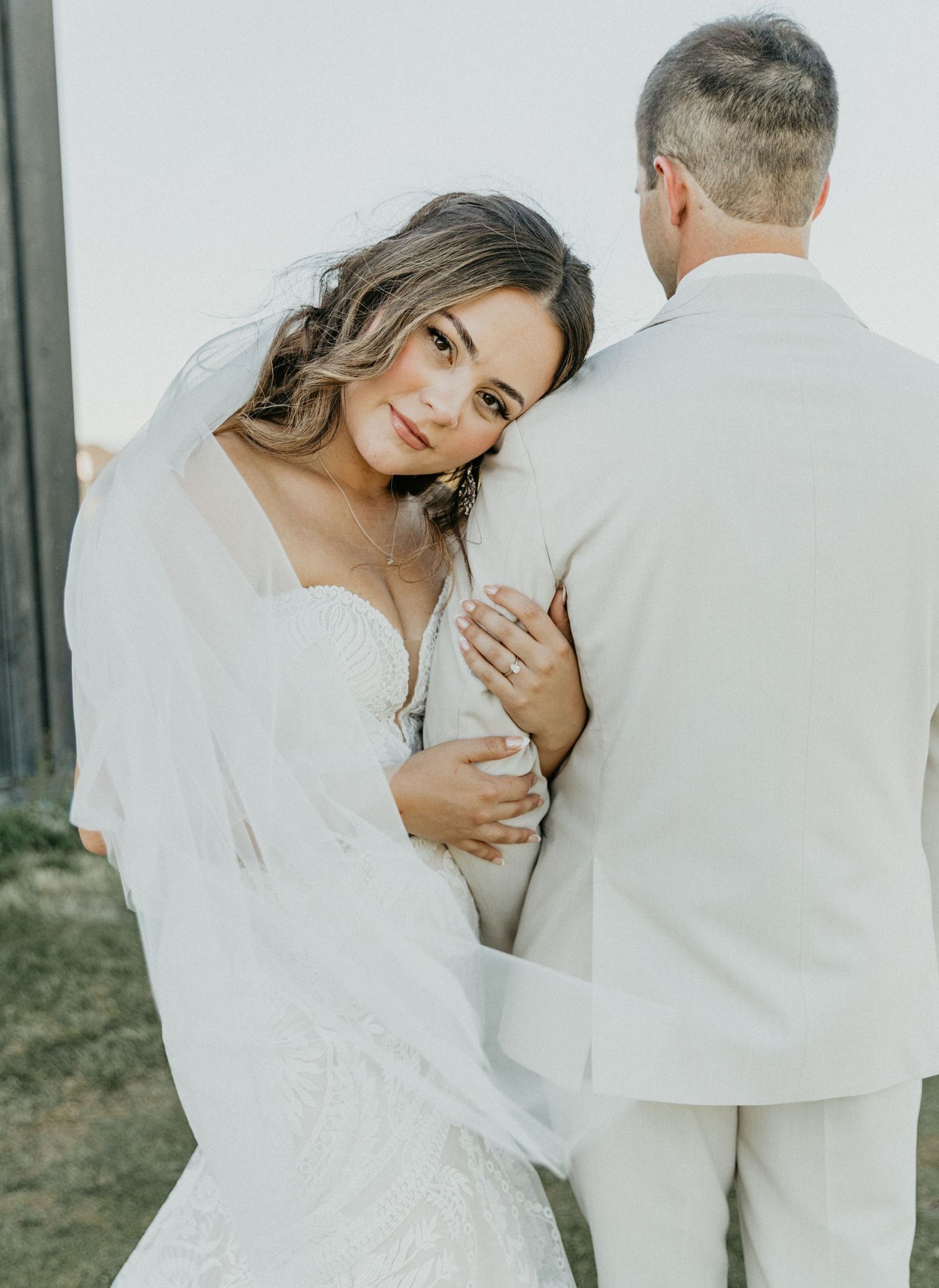 Bride in white lace dress poses intimately with groom in light suit during wedding photoshoot.