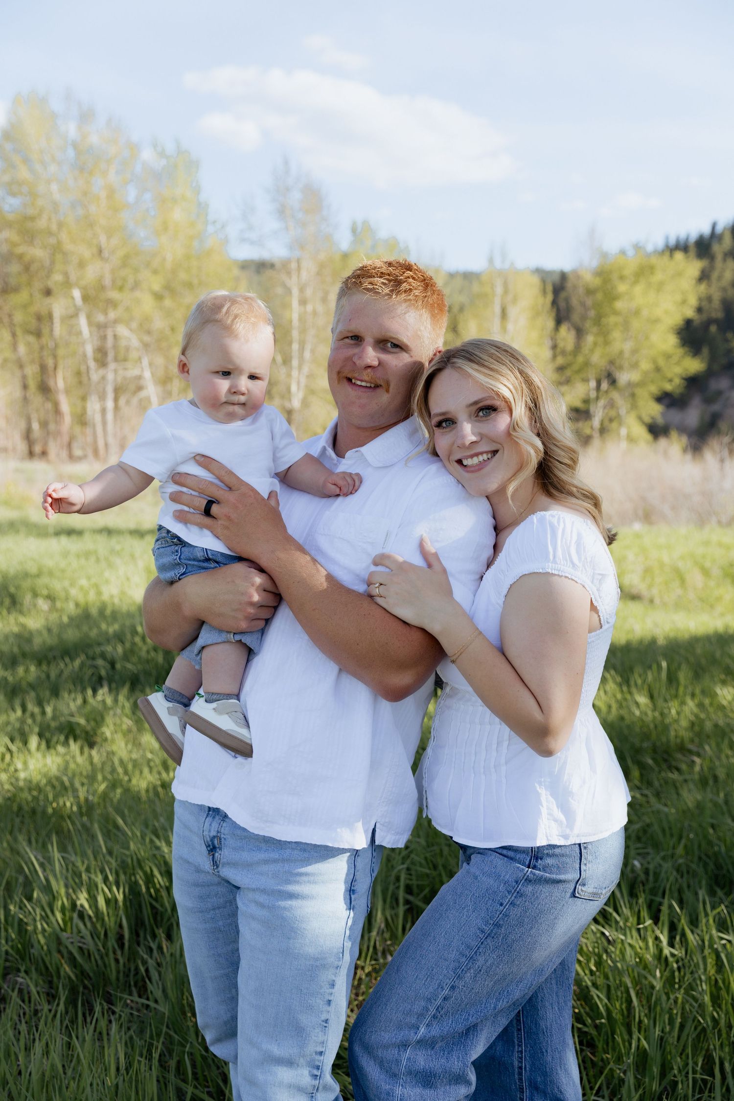 A happy family with a red-haired father, blonde mother, and baby pose in a sunny green field.