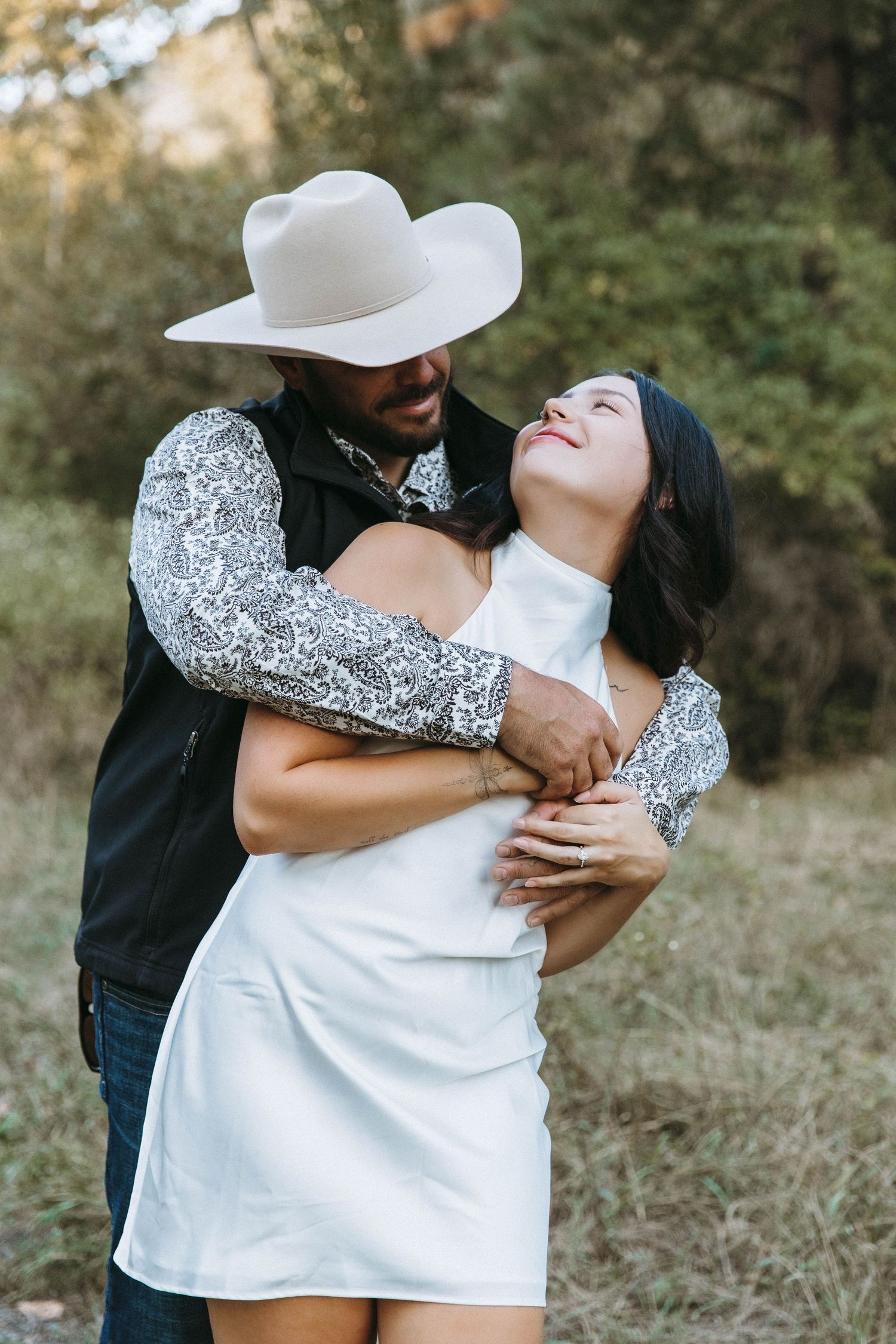 A joyful couple sharing a tender moment outdoors, with one wearing a white cowboy hat and sparkly western shirt.