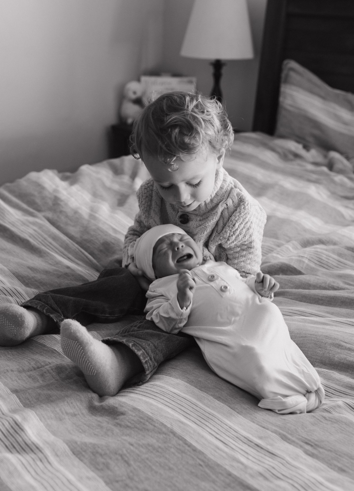 Sibling tenderly holding newborn baby on bed in black and white photo.