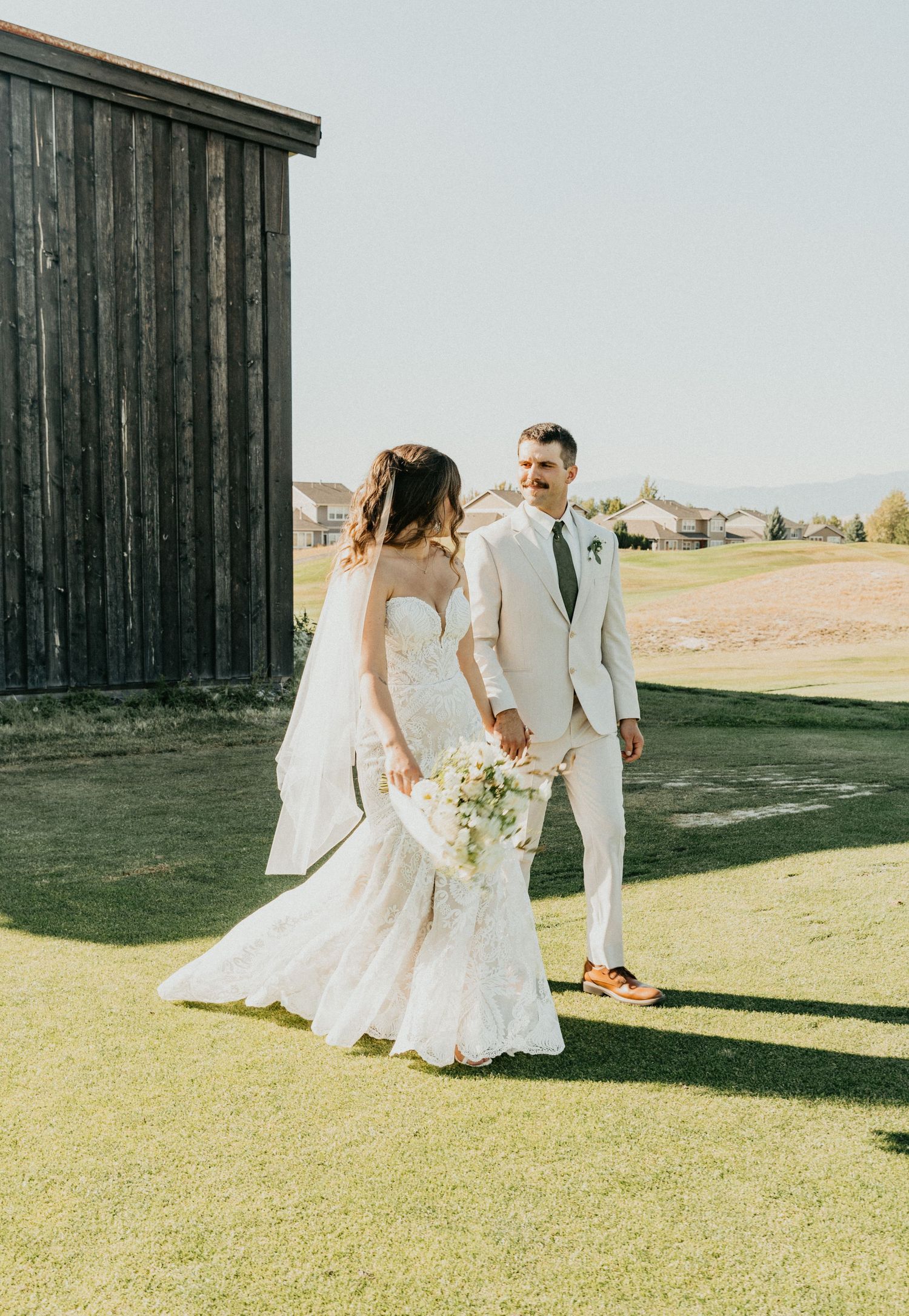 A bride and groom walking together on a grassy field next to a rustic barn on their wedding day.