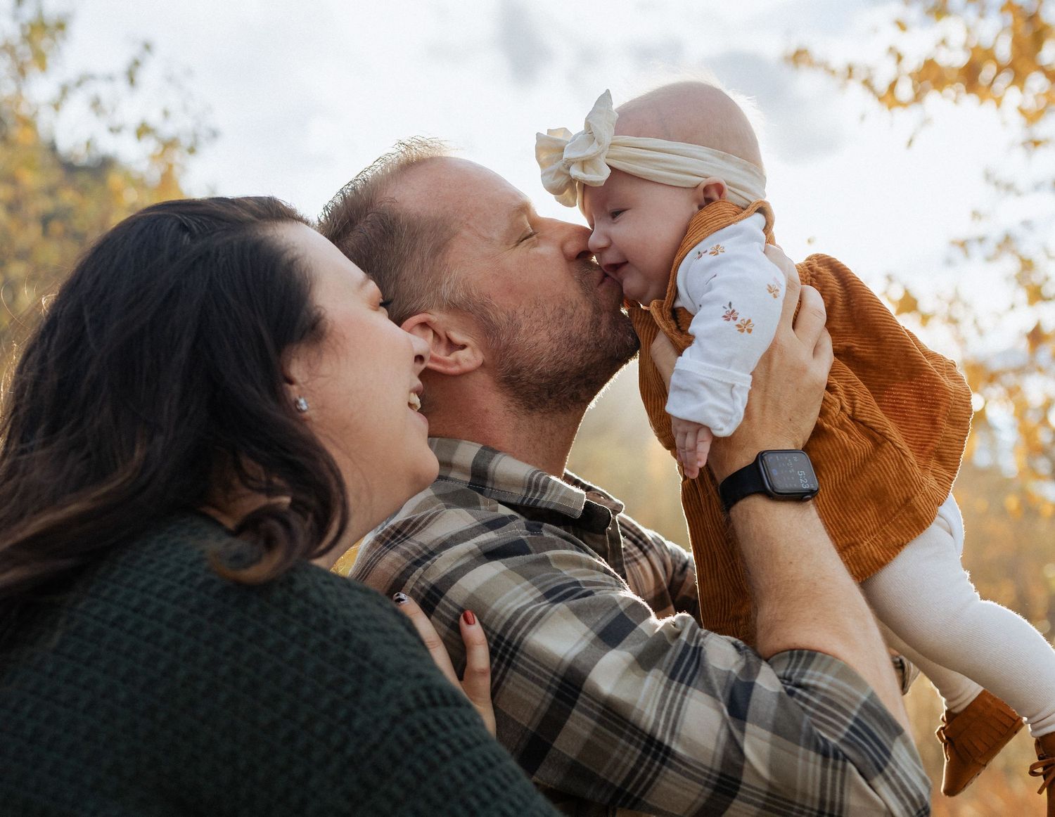 A tender family moment captured outdoors in autumn with a baby in brown overalls being lifted and kissed.