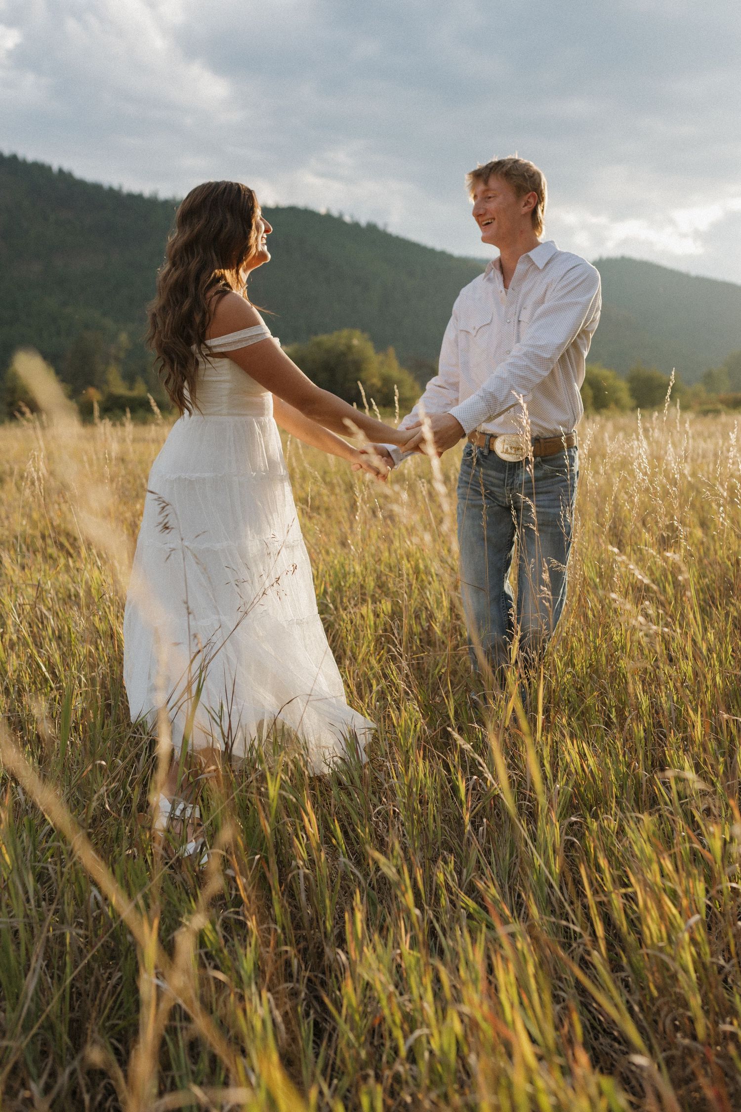 Couple holding hands while standing in tall grass with mountains in background.