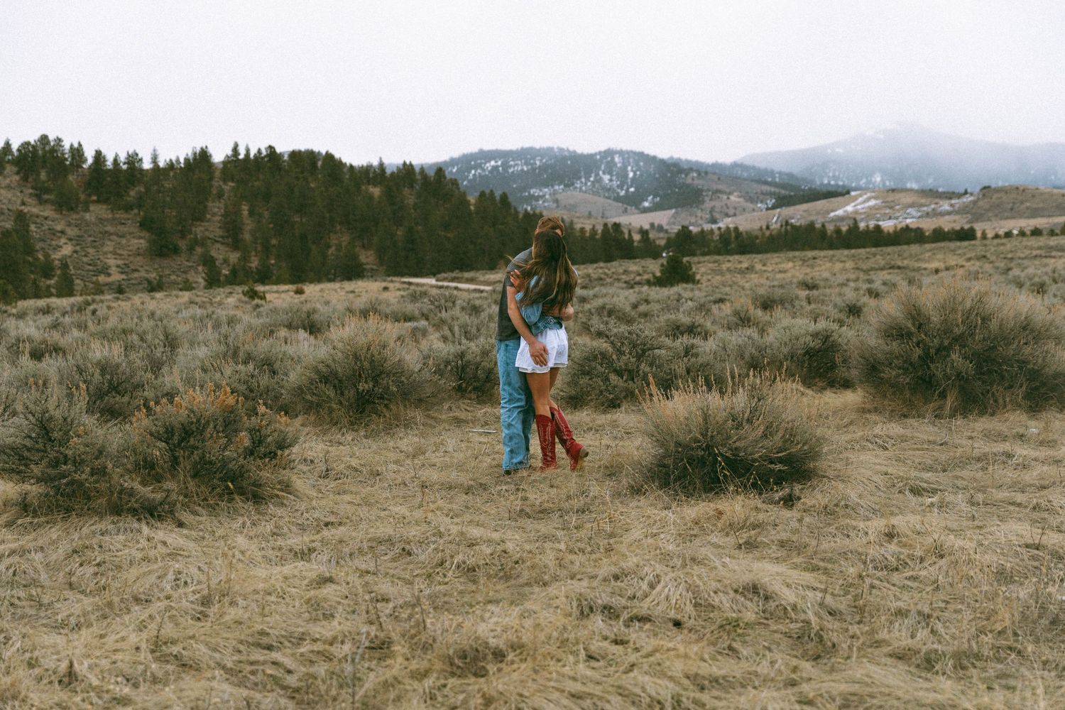 A romantic couple embraces in an open field with mountains and pine trees in the background.