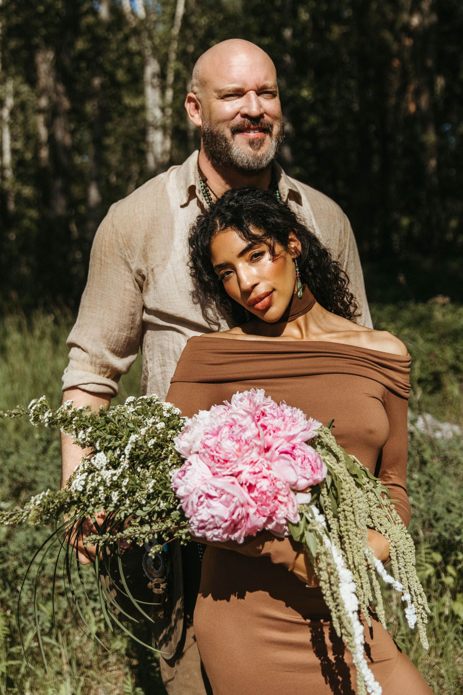 A couple in a wooded setting poses with a bouquet of pink flowers and greenery in a romantic outdoor photoshoot.