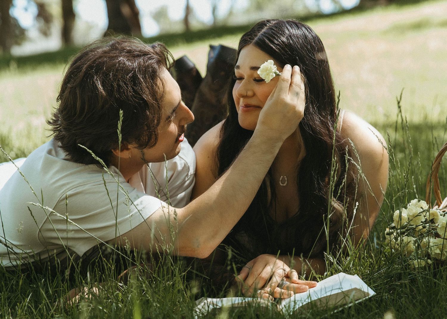 A couple lies in a grassy meadow on a sunny day, sharing an intimate moment together outdoors.