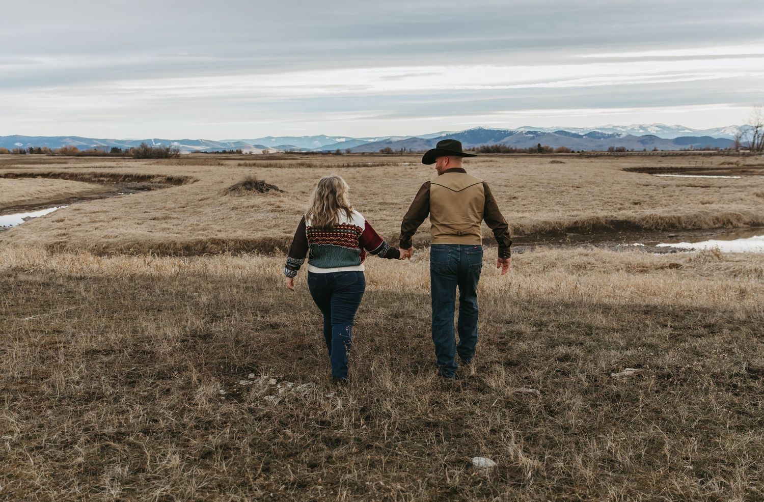 Two people walk through a brown winter field with mountains visible in the distance on an overcast day.