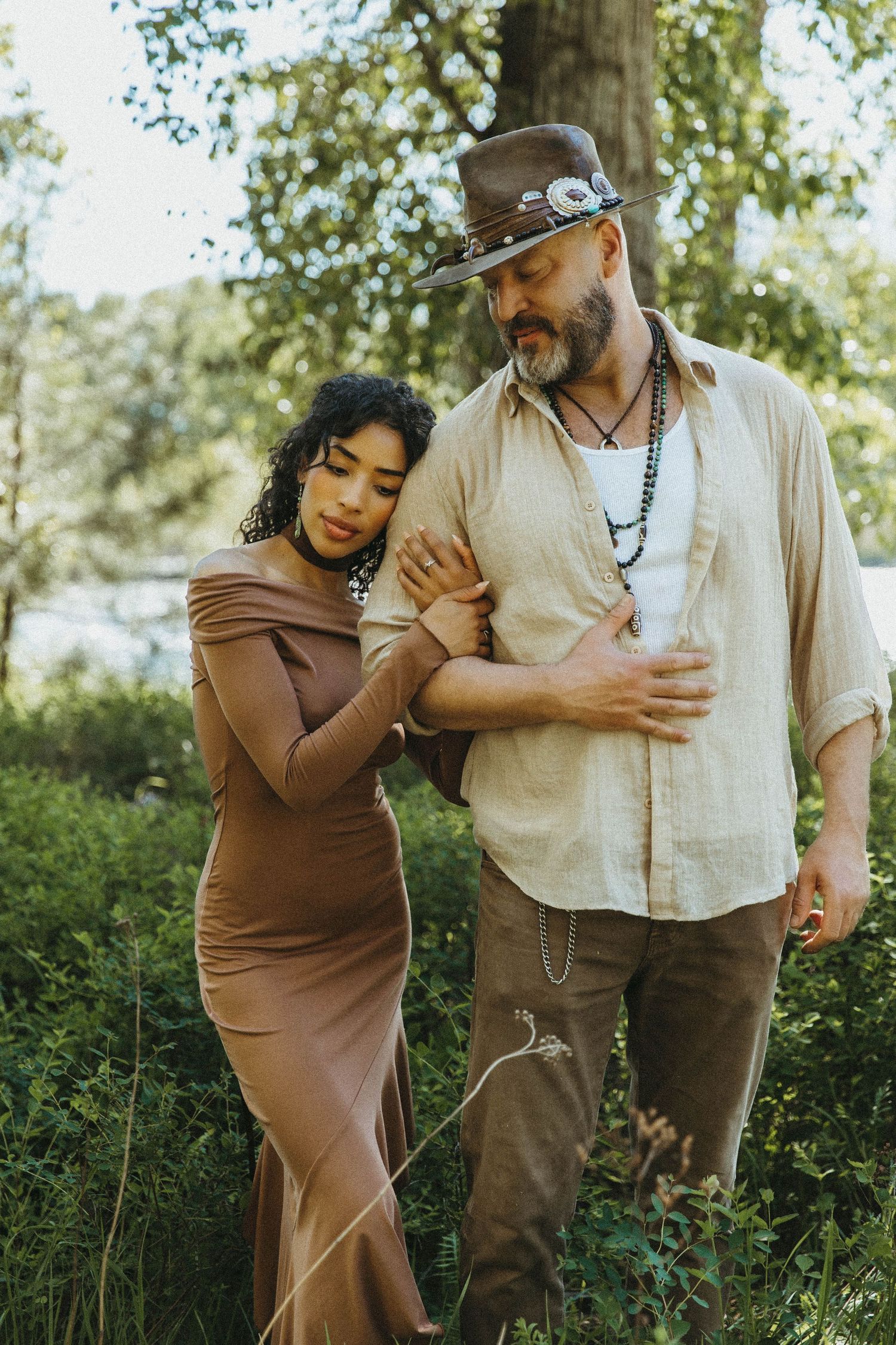 A couple in casual attire embraces tenderly in a sun-dappled garden setting with green foliage in the background.