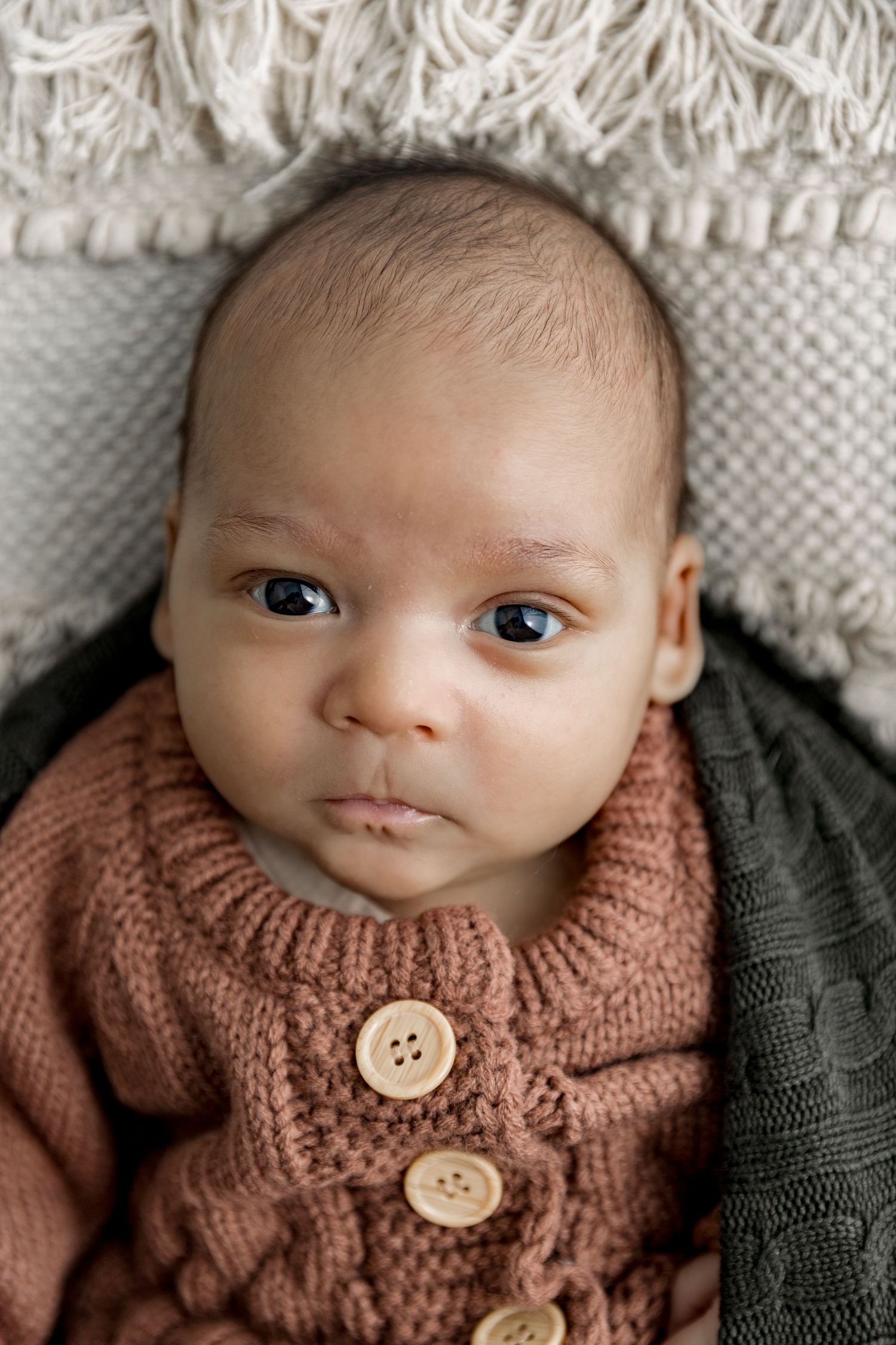 Baby wearing cozy knit sweater with wooden buttons lies on textured white blanket looking at camera.