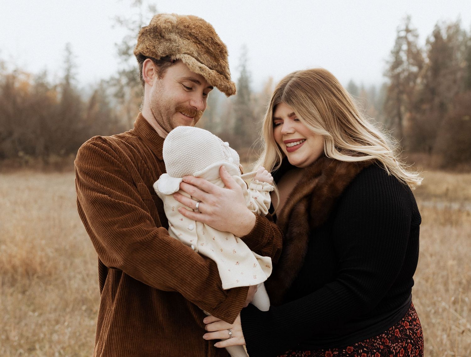Couple in warm fall clothing holding a small baby in a peaceful autumn field setting.