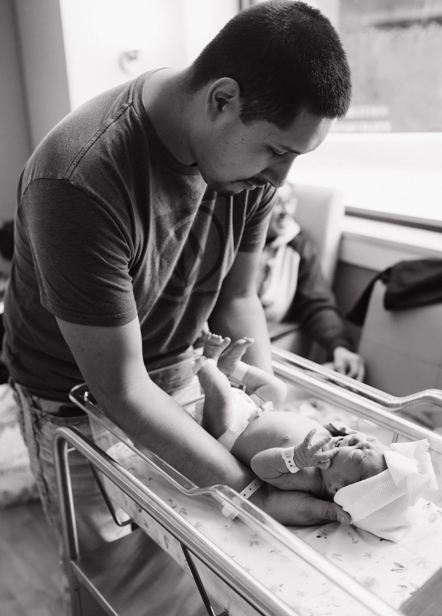 A black and white photograph shows someone tenderly caring for a baby on bedside bassinet.