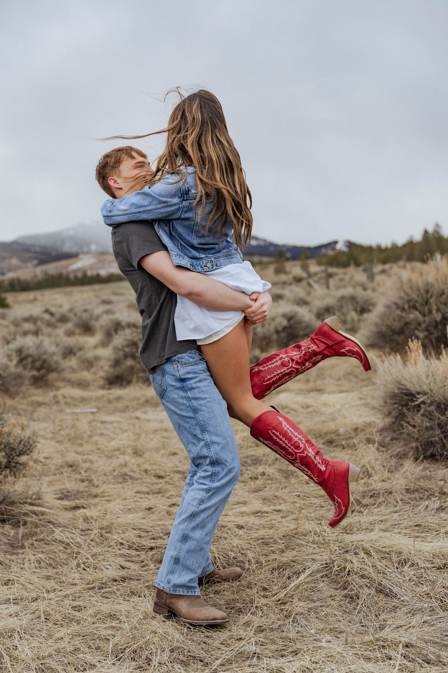 A playful couple shares a joyful moment wearing denim and red boots against a mountain landscape.