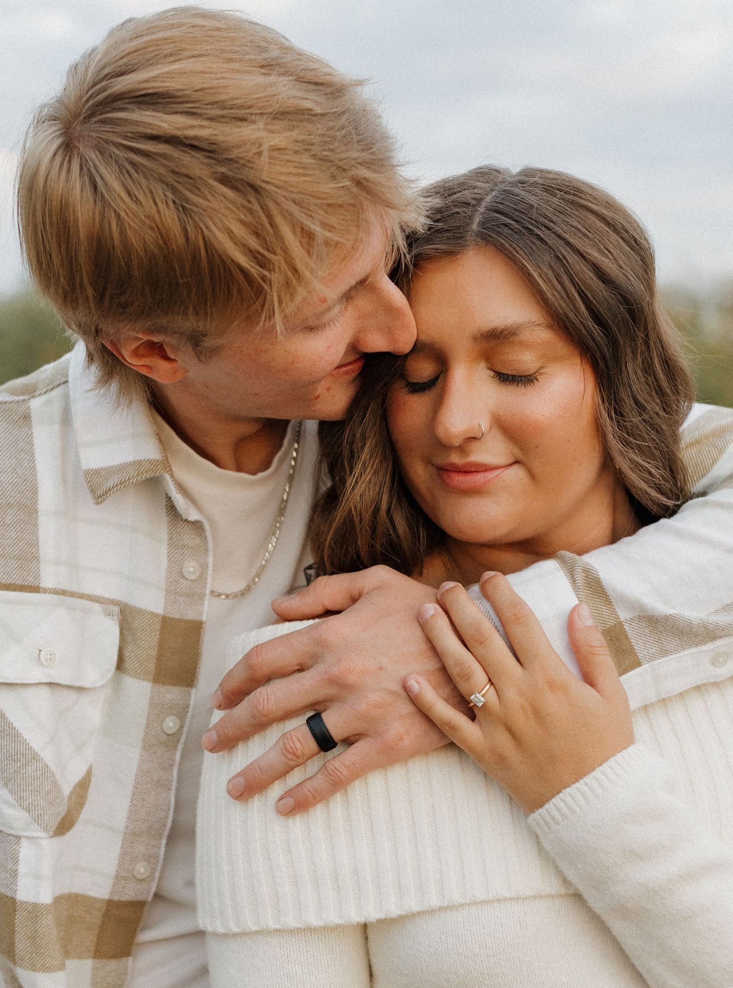 Couple in cozy white sweaters embracing while showing engagement ring.