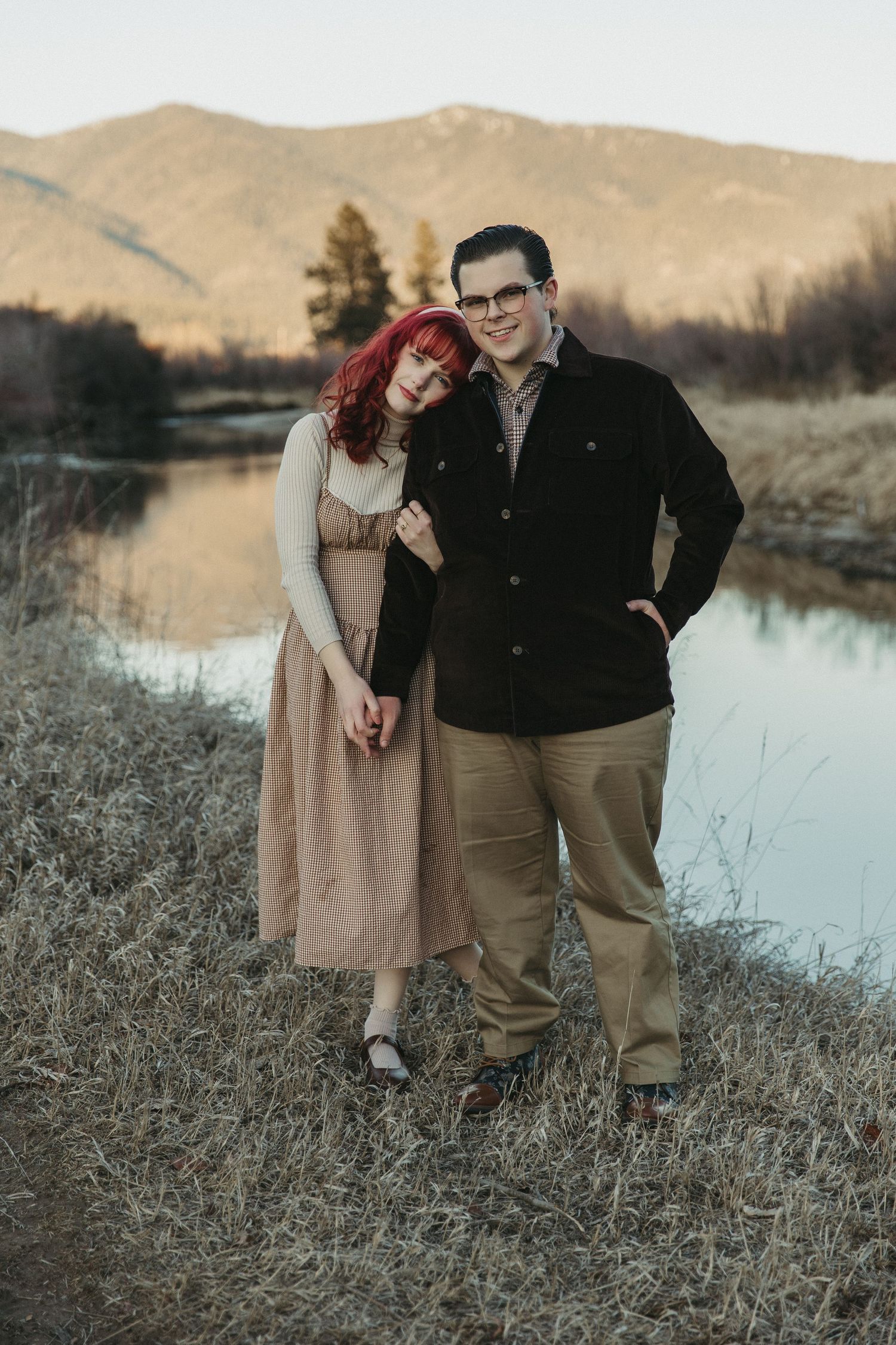 A couple stands together by a tranquil lake at sunset with mountains in the background on a peaceful autumn evening.
