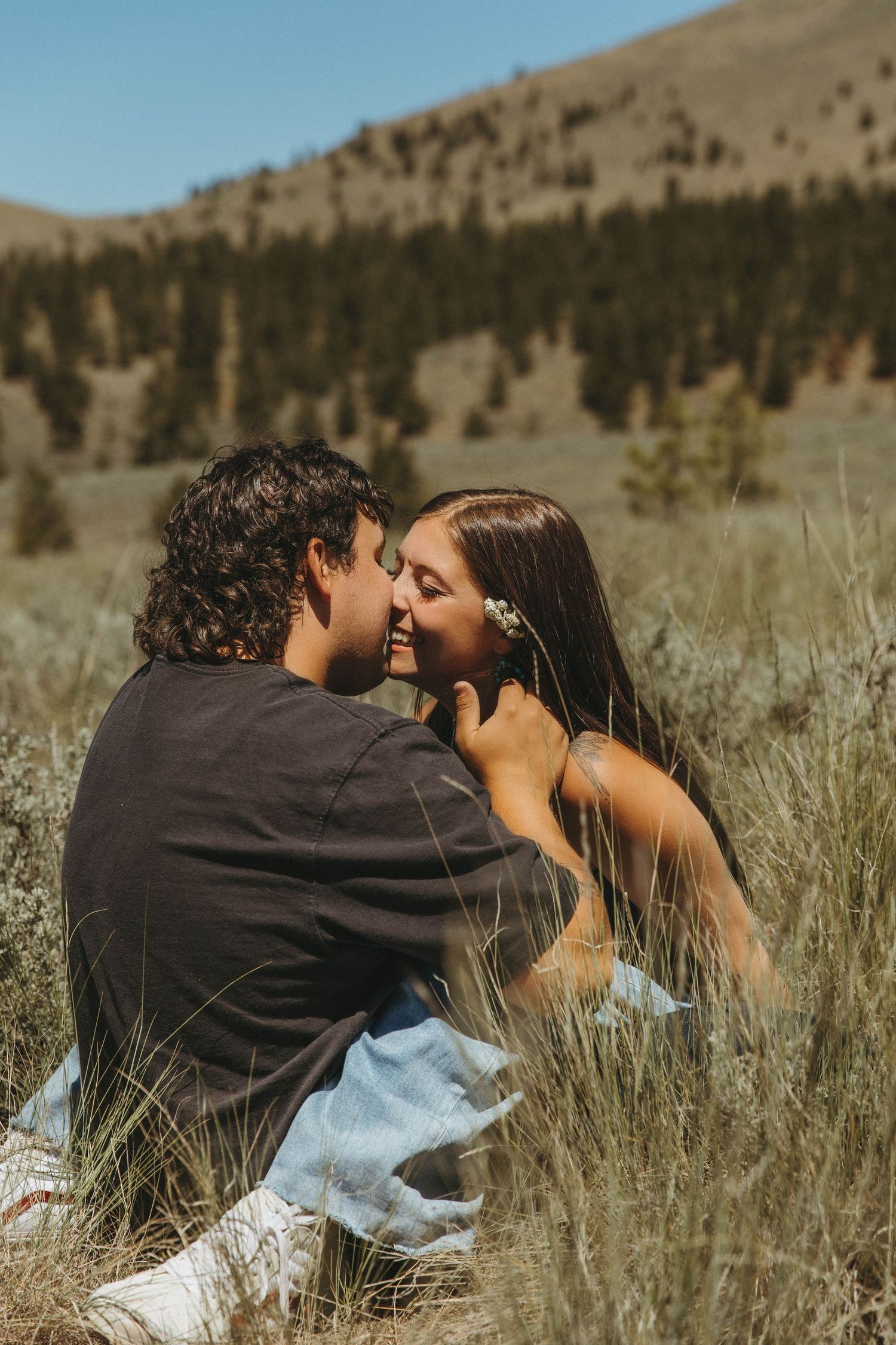 A romantic couple sharing a tender moment while sitting in tall grass with mountains and forest in the background.