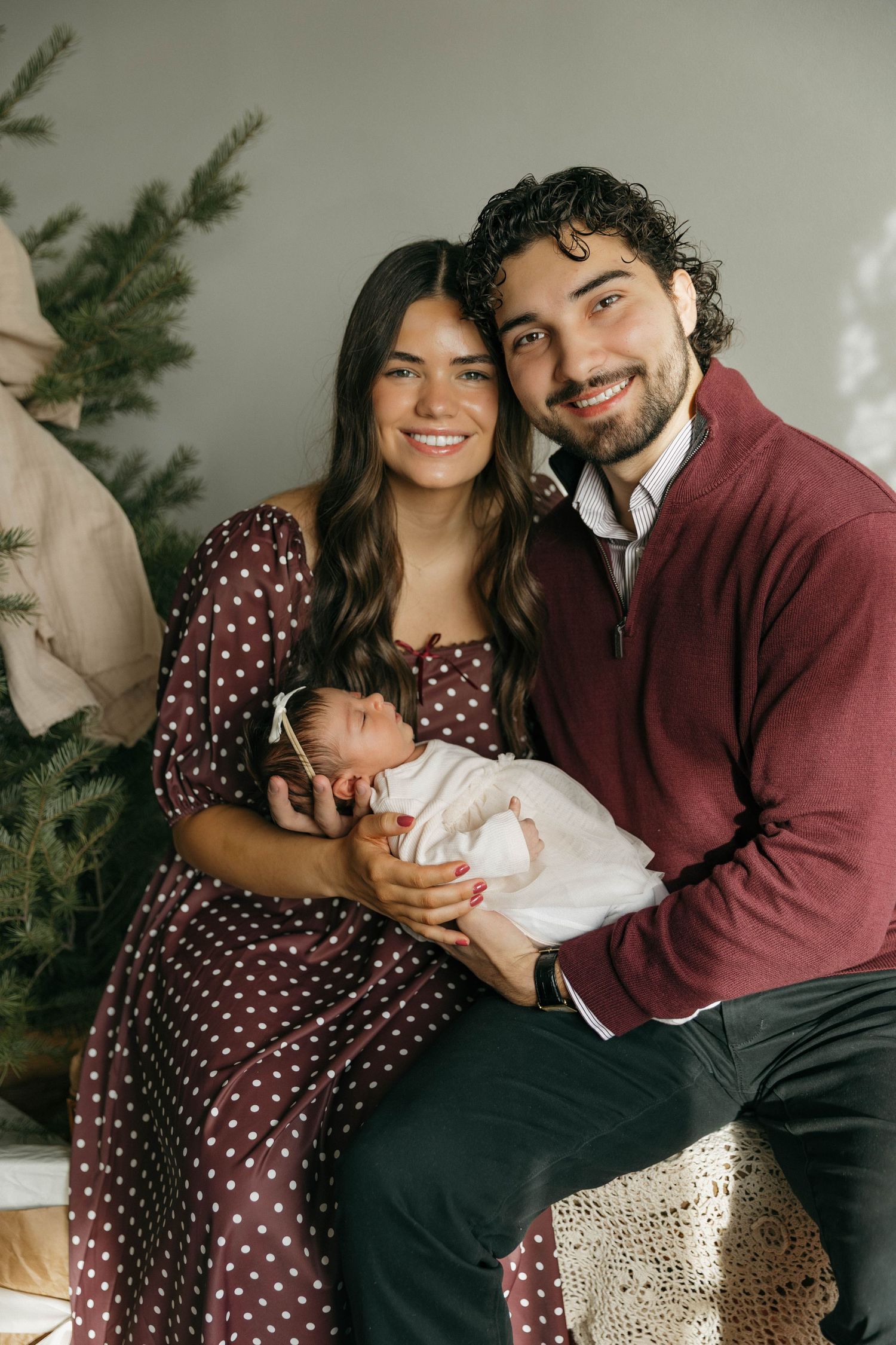 A couple in burgundy and polka dots share tender moments with their newborn baby on a couch with holiday decor.