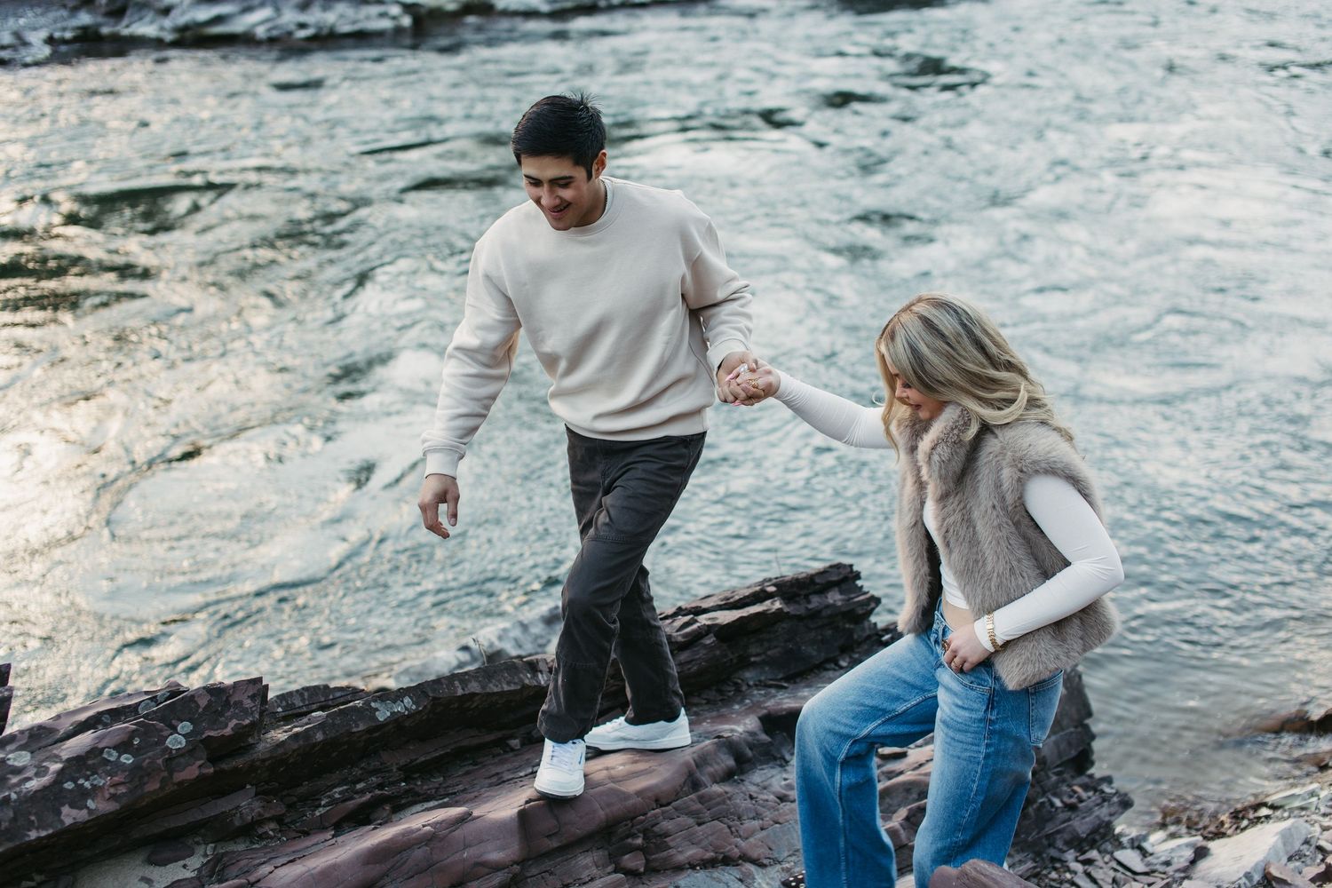 Two people walking together along rocky coastal terrain near ocean waves on an overcast day.