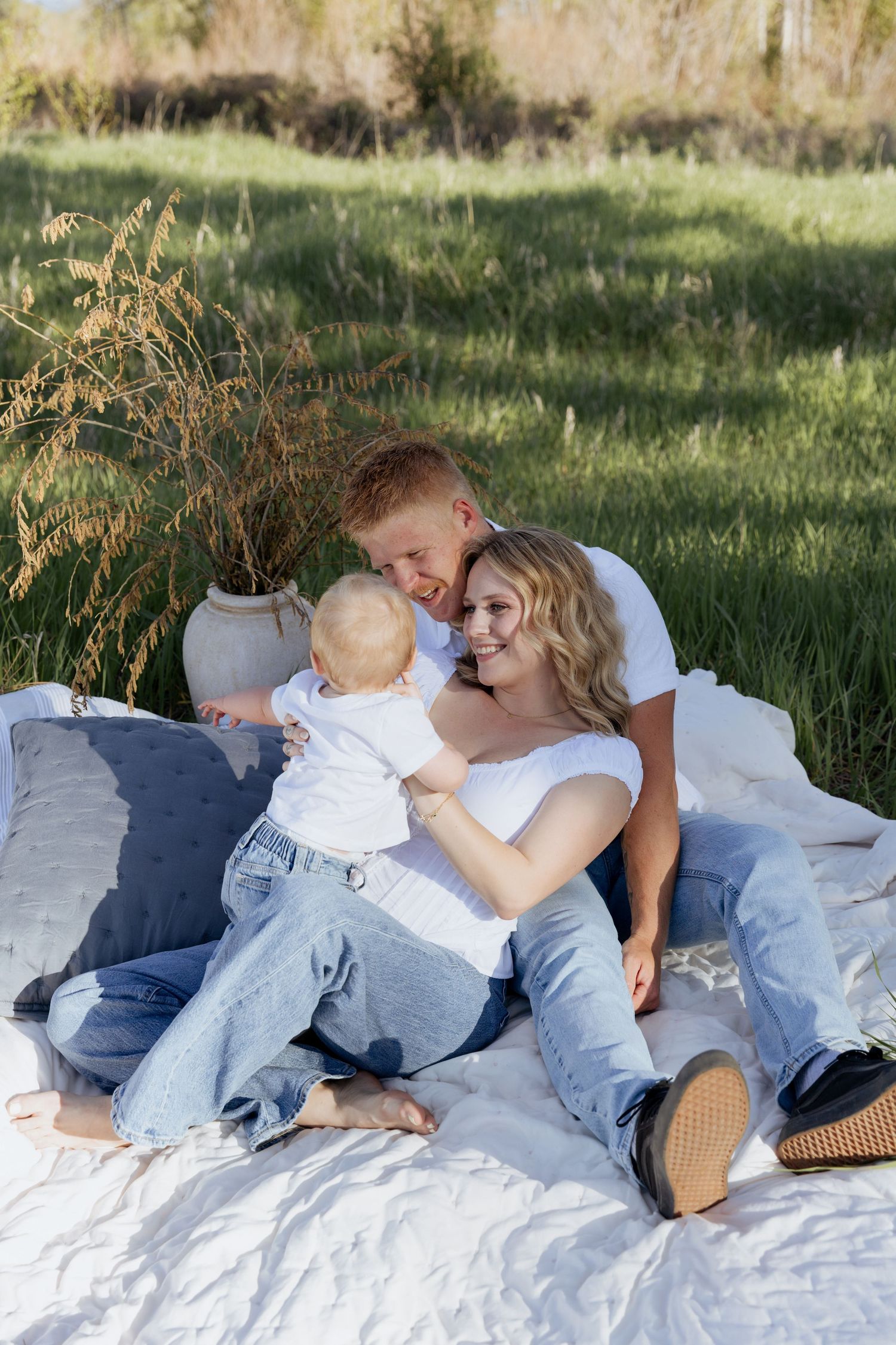 Young family with baby enjoying outdoor picnic on white blanket in green meadow, wearing matching white tops and jeans.