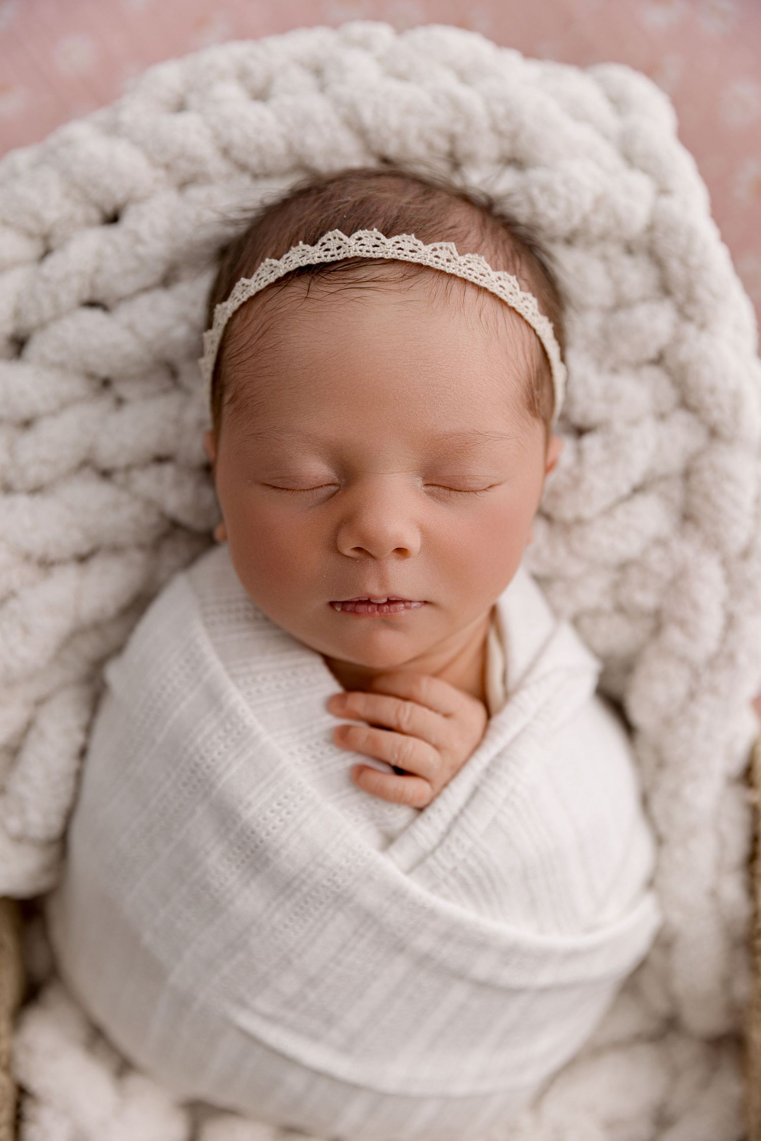 Sleeping newborn baby girl swaddled in white wrap with lace headband on chunky knit blanket.
