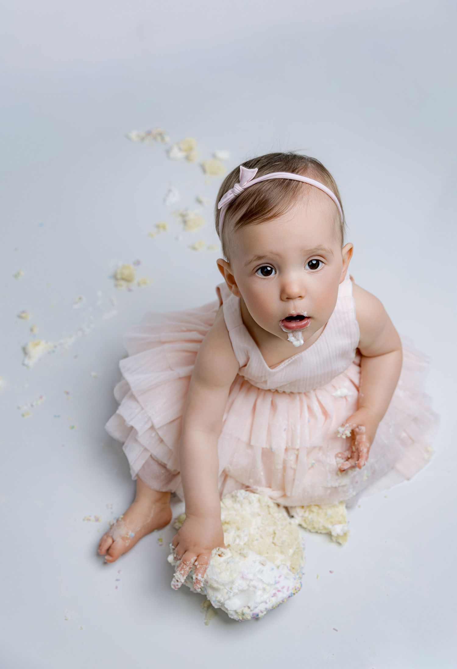 Baby girl in pink tutu dress smashes first birthday cake, sitting on light blue backdrop with frosting on her face.