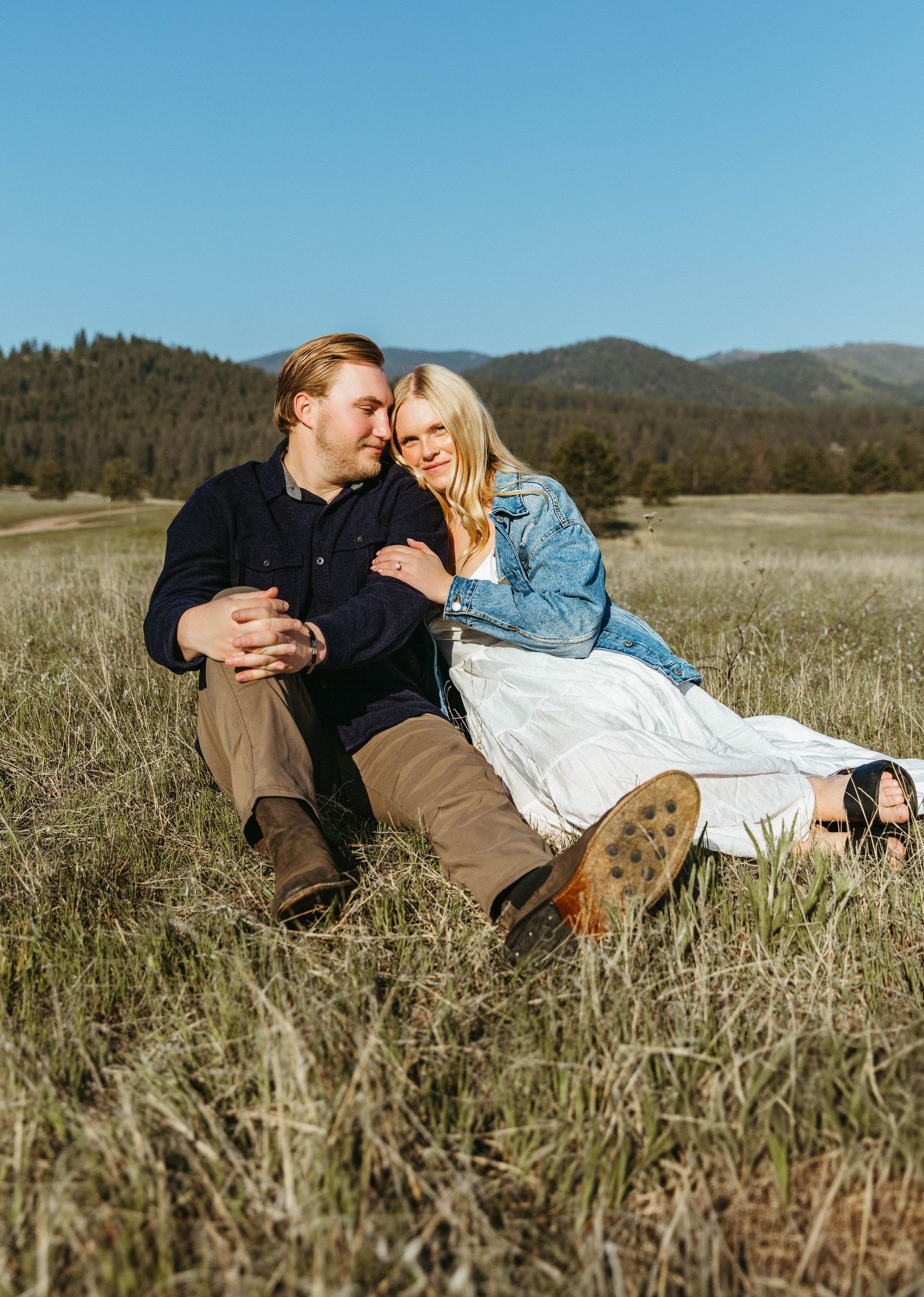 Couple lying together in a rustic grassy meadow with mountains in the background on a sunny day.