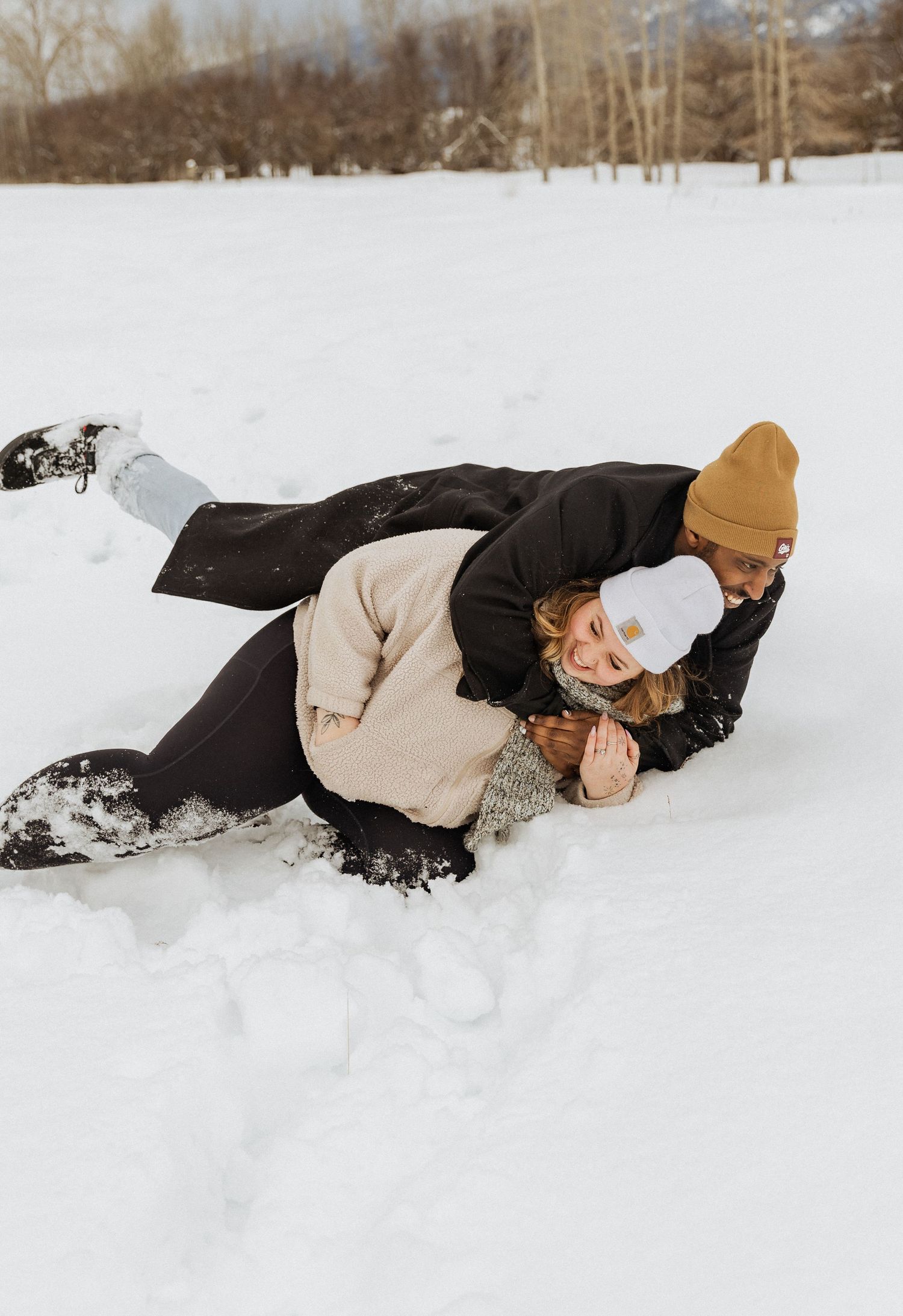 Couple playing in the snow as one person lies down wearing a beige sweater and mustard beanie.