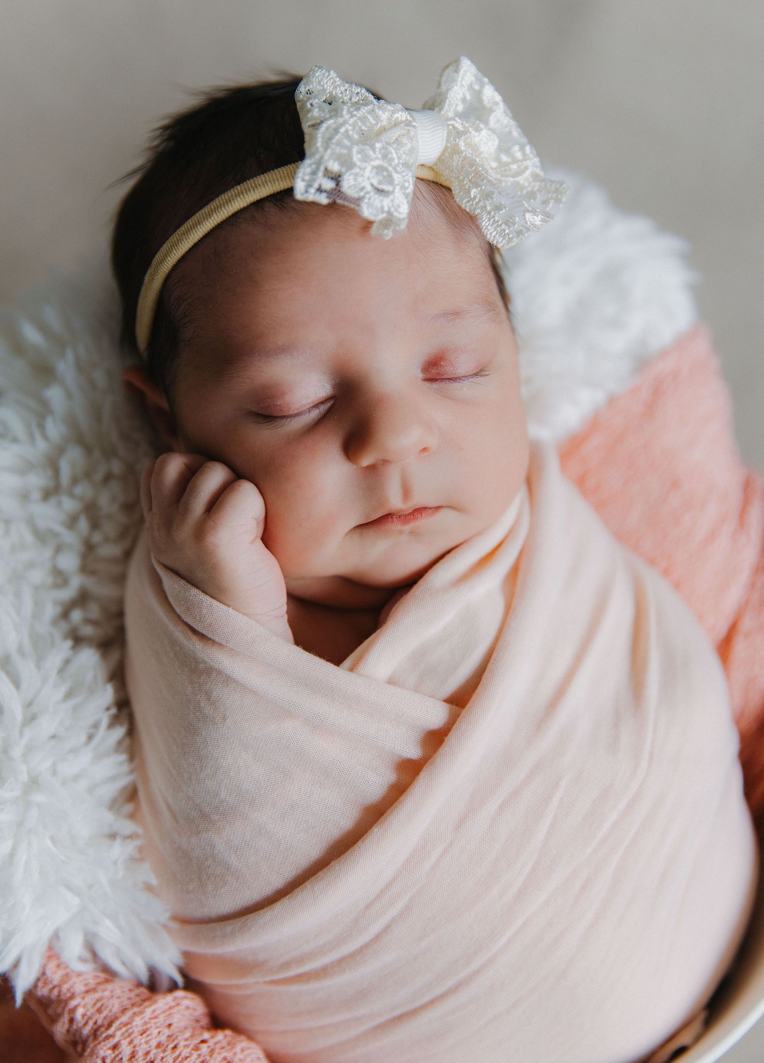 Sleeping newborn wrapped in pink swaddle with lace bow on white fur background.