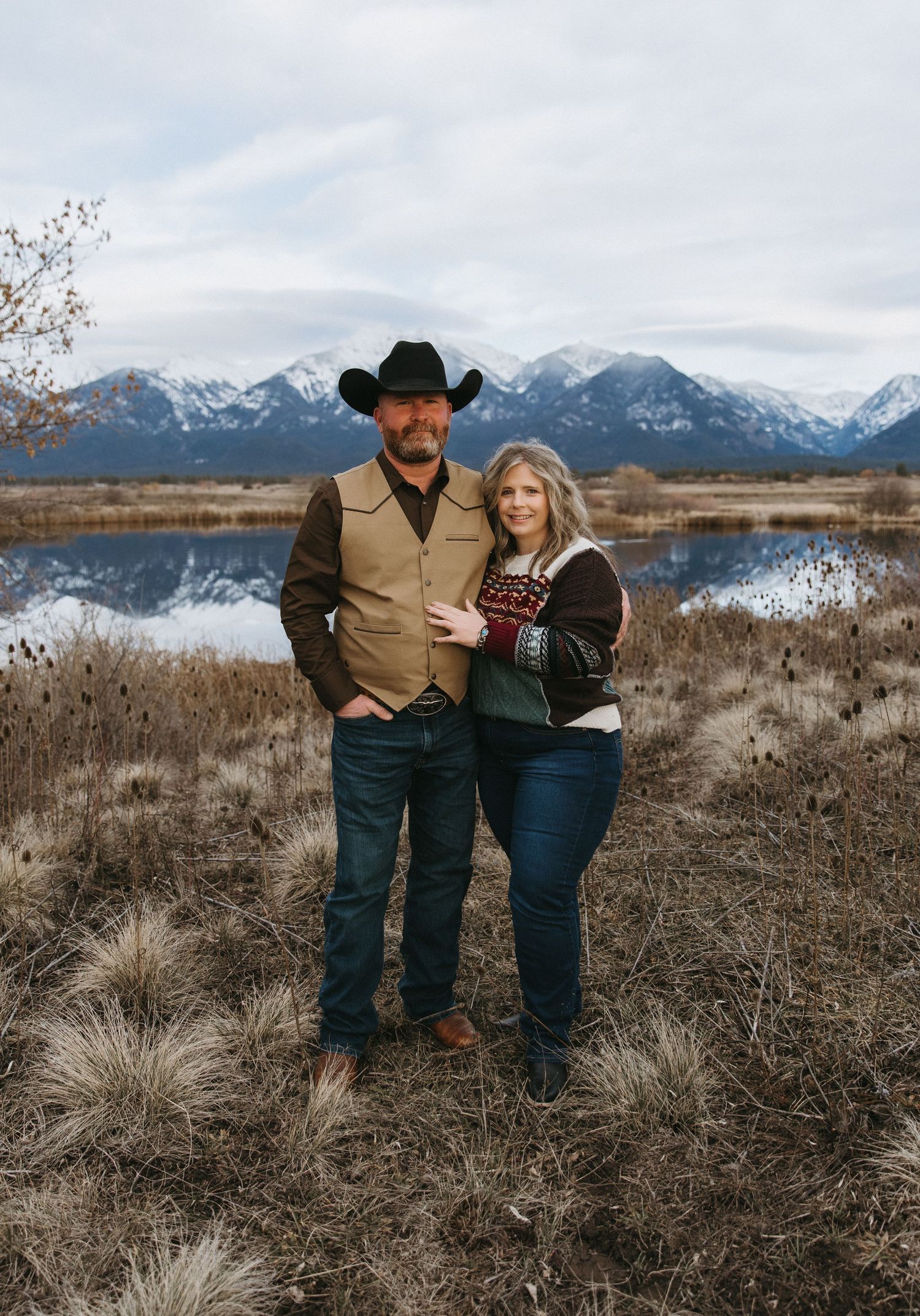 Romantic couple poses together against snowy mountain backdrop in rustic western attire.