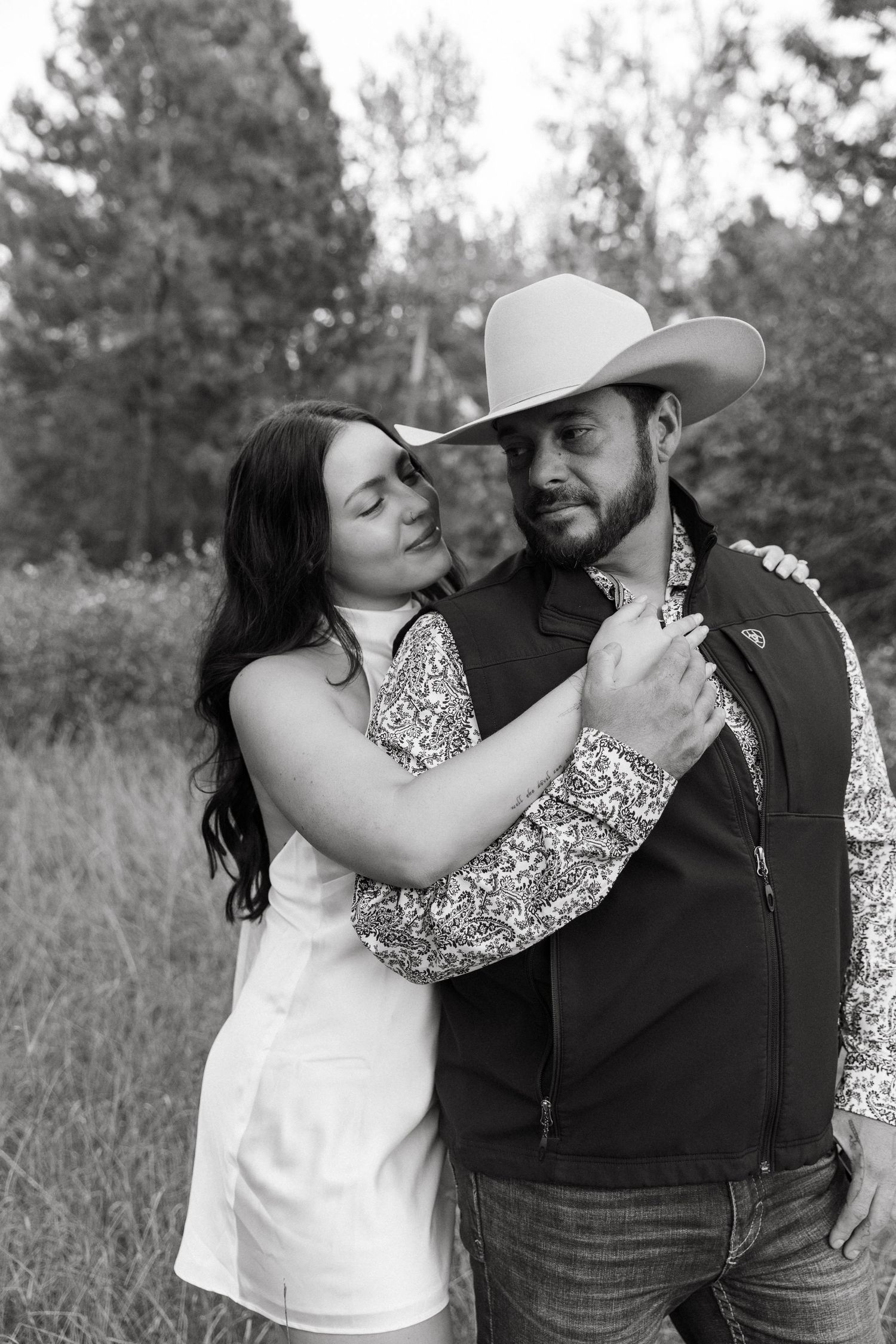 A black and white portrait of a couple in western attire embracing in a rustic outdoor setting.