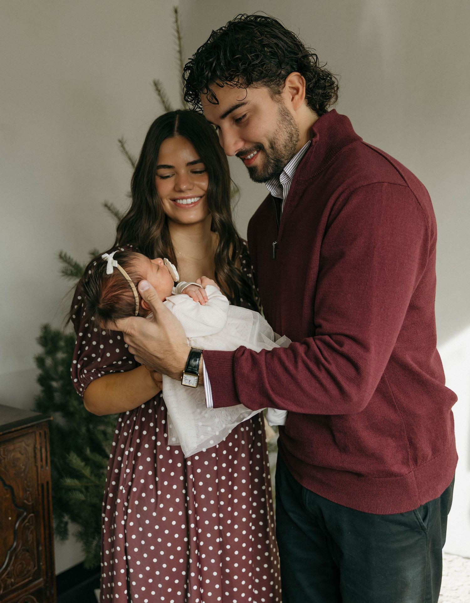 A couple in burgundy and polka dot outfits share a tender moment while holding a baby in white clothing.