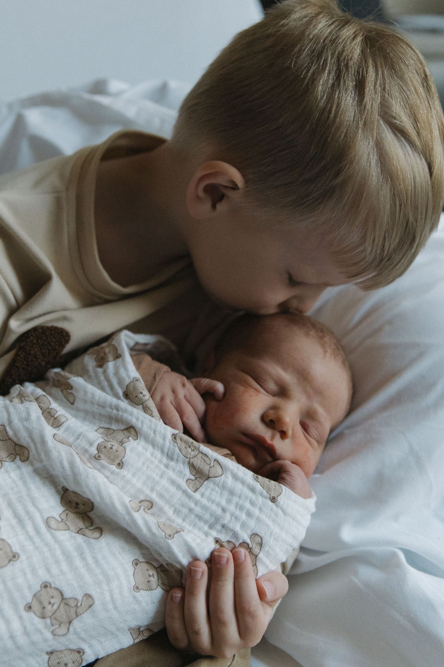 Young boy gently kisses sleeping newborn baby wrapped in a white teddy bear muslin swaddle blanket.