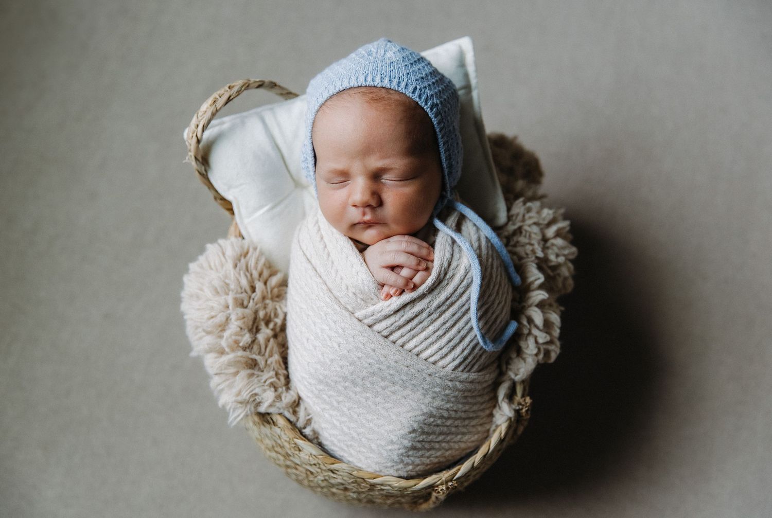 Newborn baby swaddled in white wrap with blue hat sleeping peacefully in woven basket.