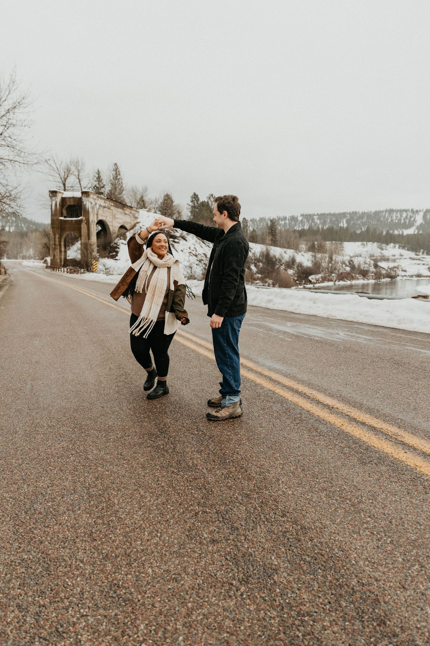 A playful couple dancing together on a rural road with snow-covered mountains in the background.