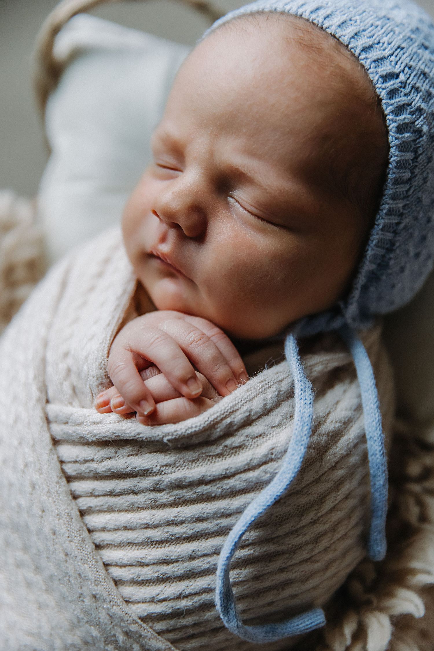 Sleeping newborn in white wrap and blue knit hat with hands folded.