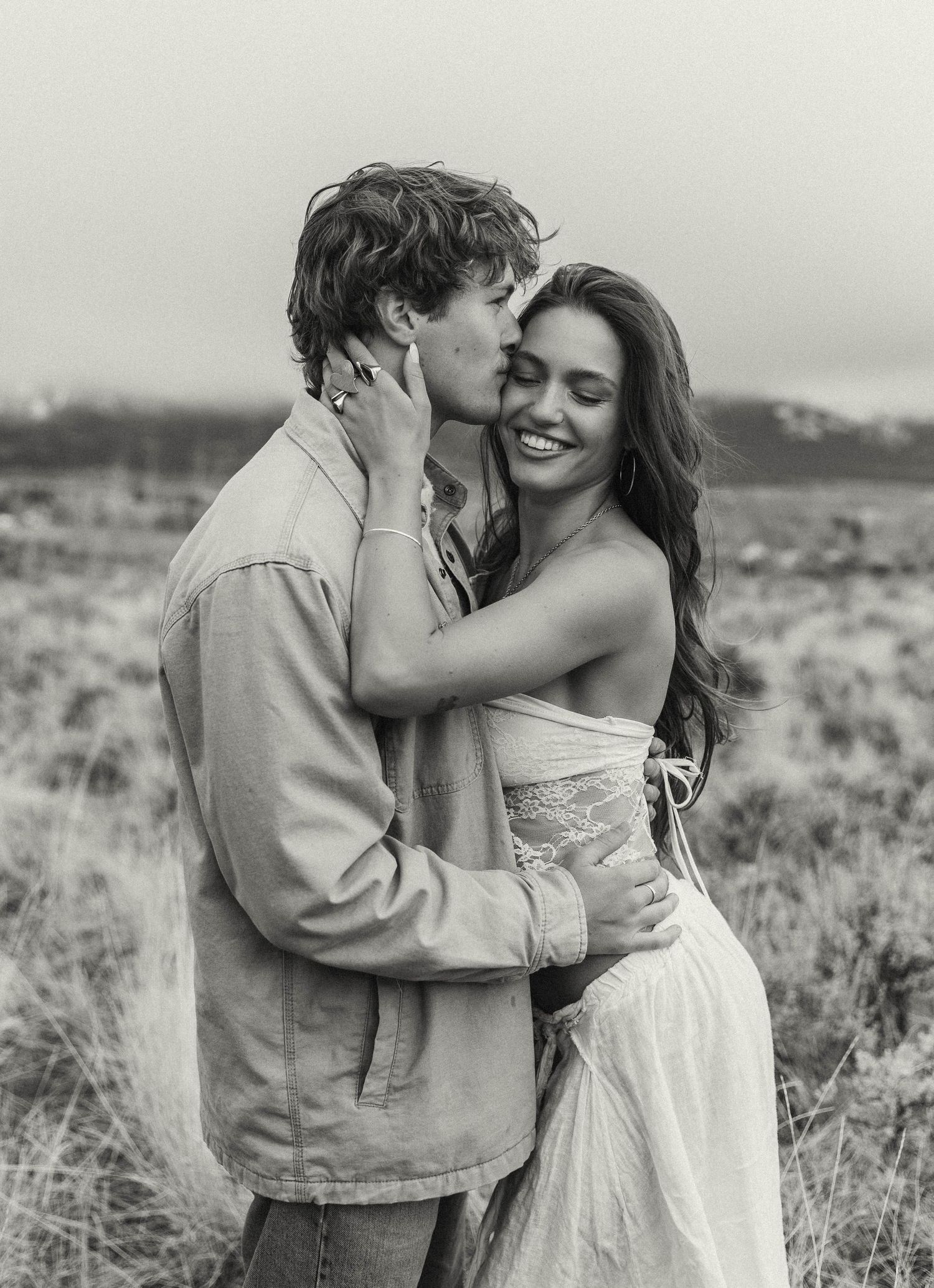 Romantic couple embracing in a field during sunset wearing casual attire.