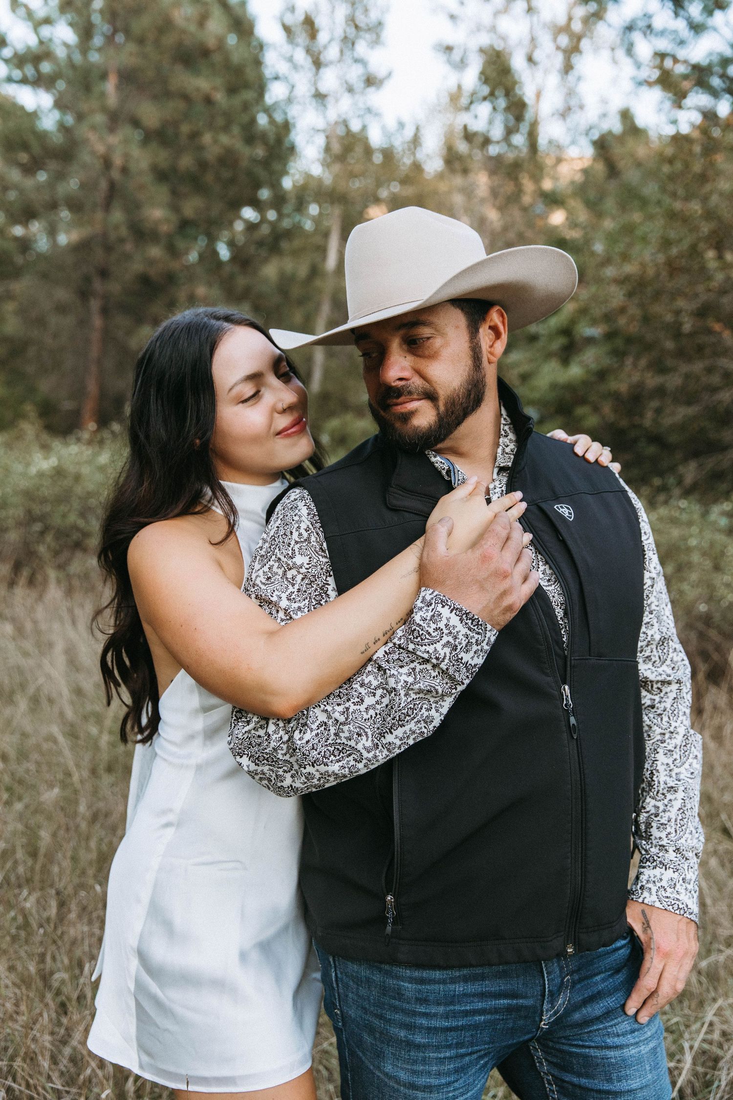 A romantic couple in western-inspired outfits pose together against a forested background.