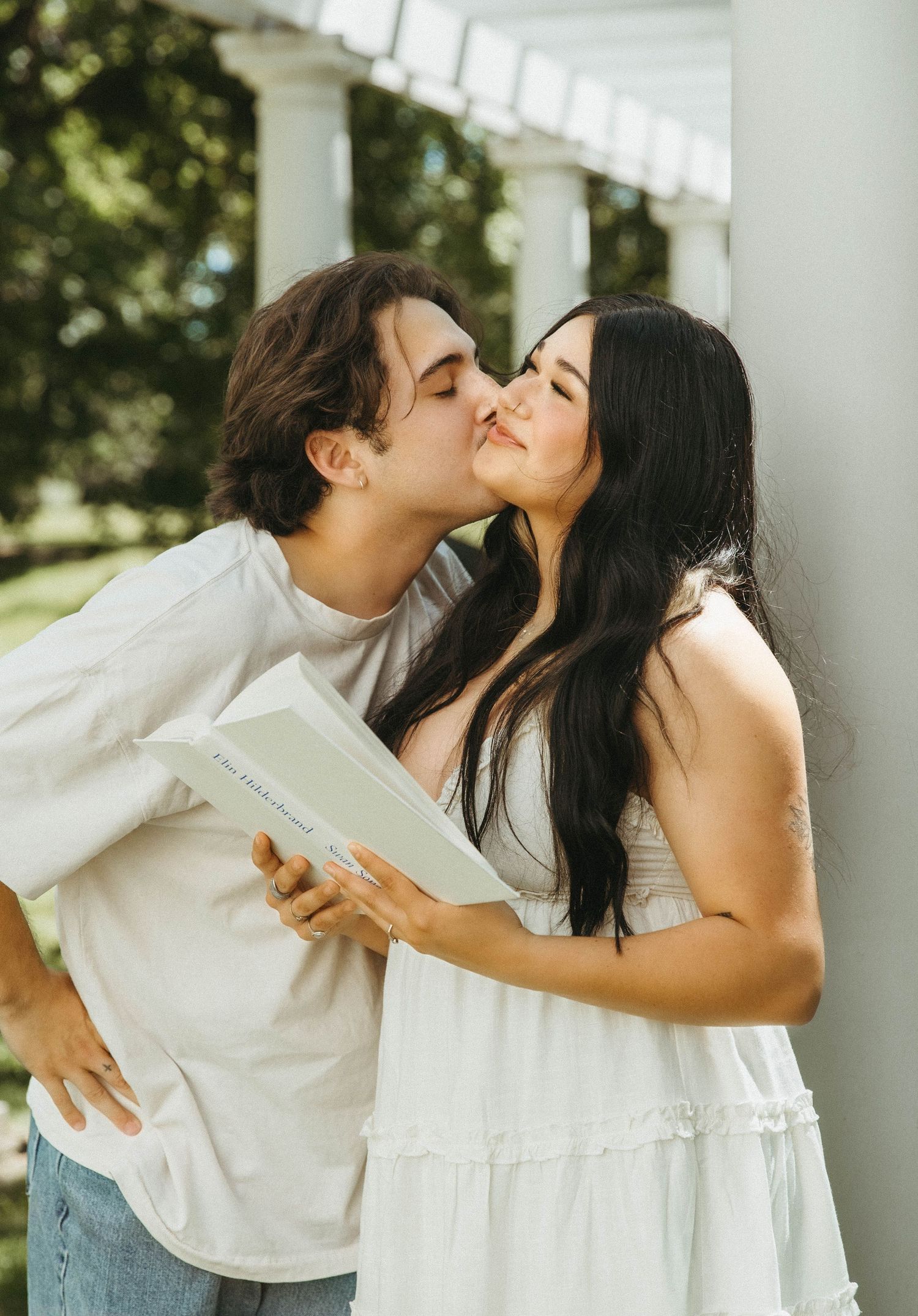 A couple in white outfits shares a series of romantic kisses while standing near a white column on a bright sunny day.